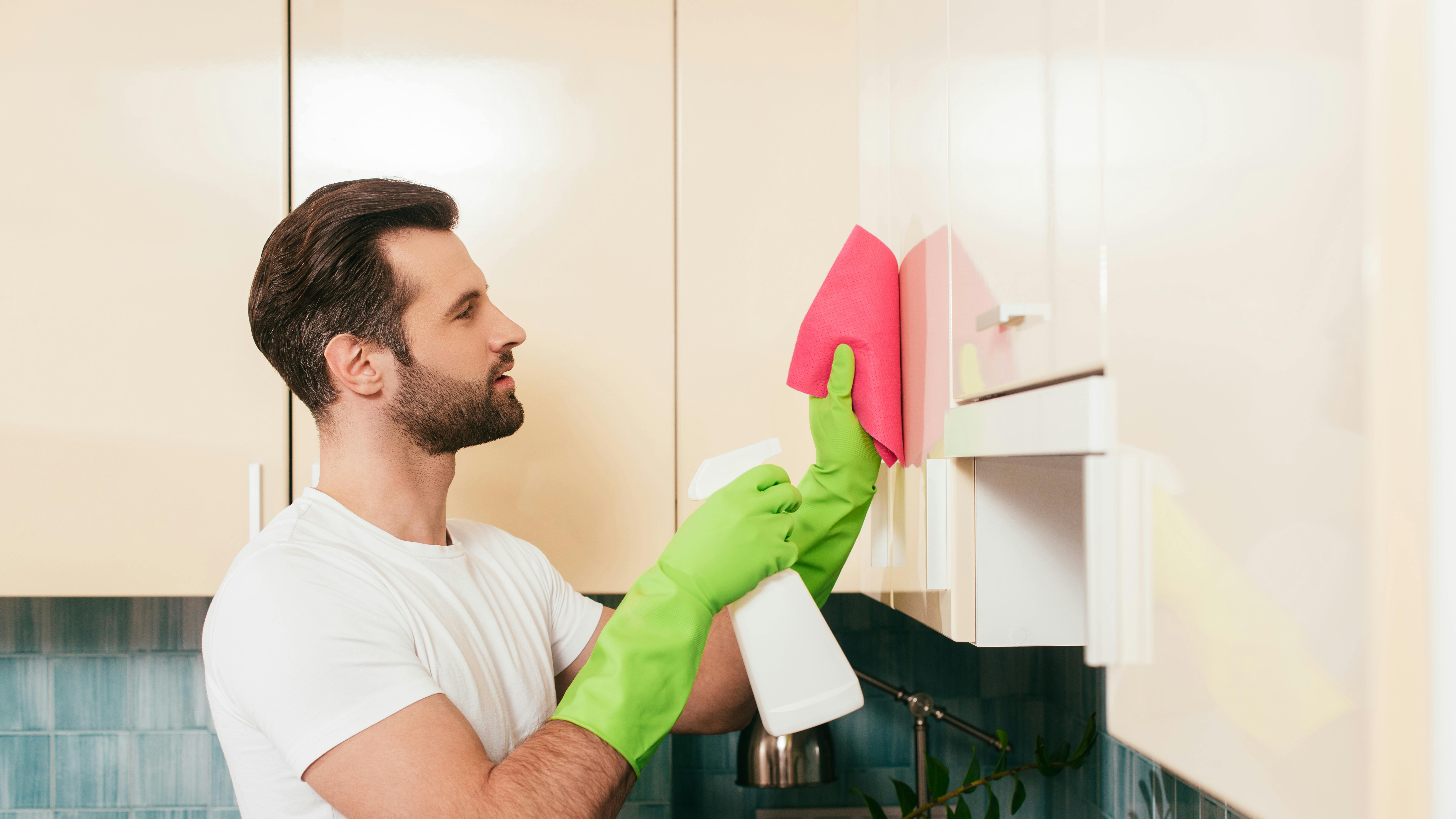 Side view of handsome man using detergent and rag while cleaning cupboard in kitchen