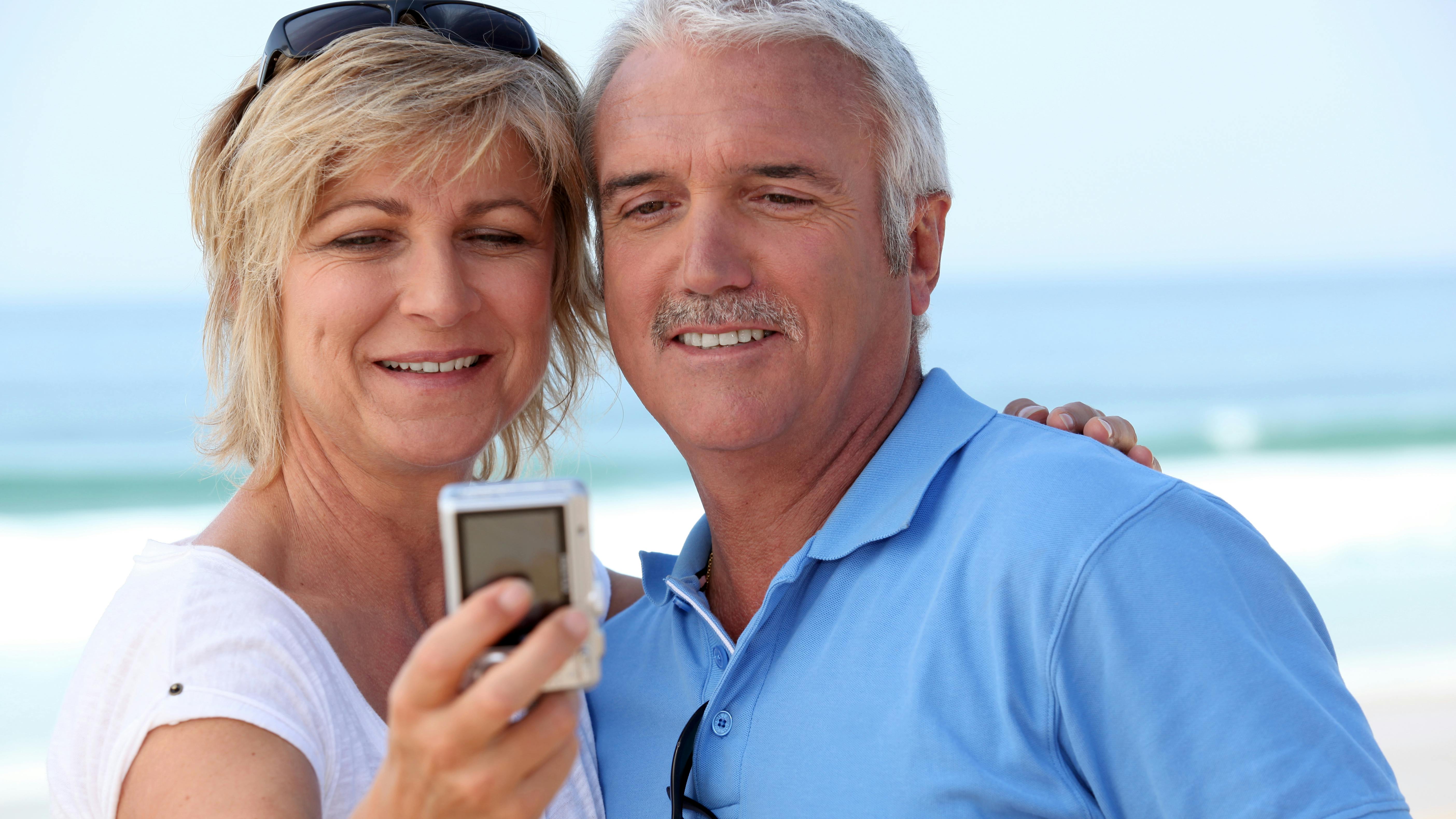 Forelsket midaldrende par tager en selfie på stranden med havet i baggrunden