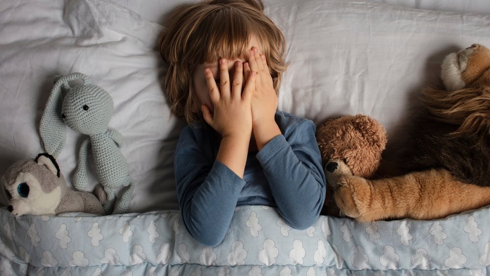 Dreng på 6-7 år ligger i sin seng og holder hænderne for øjnene. Han er omgivet at sine bamser.