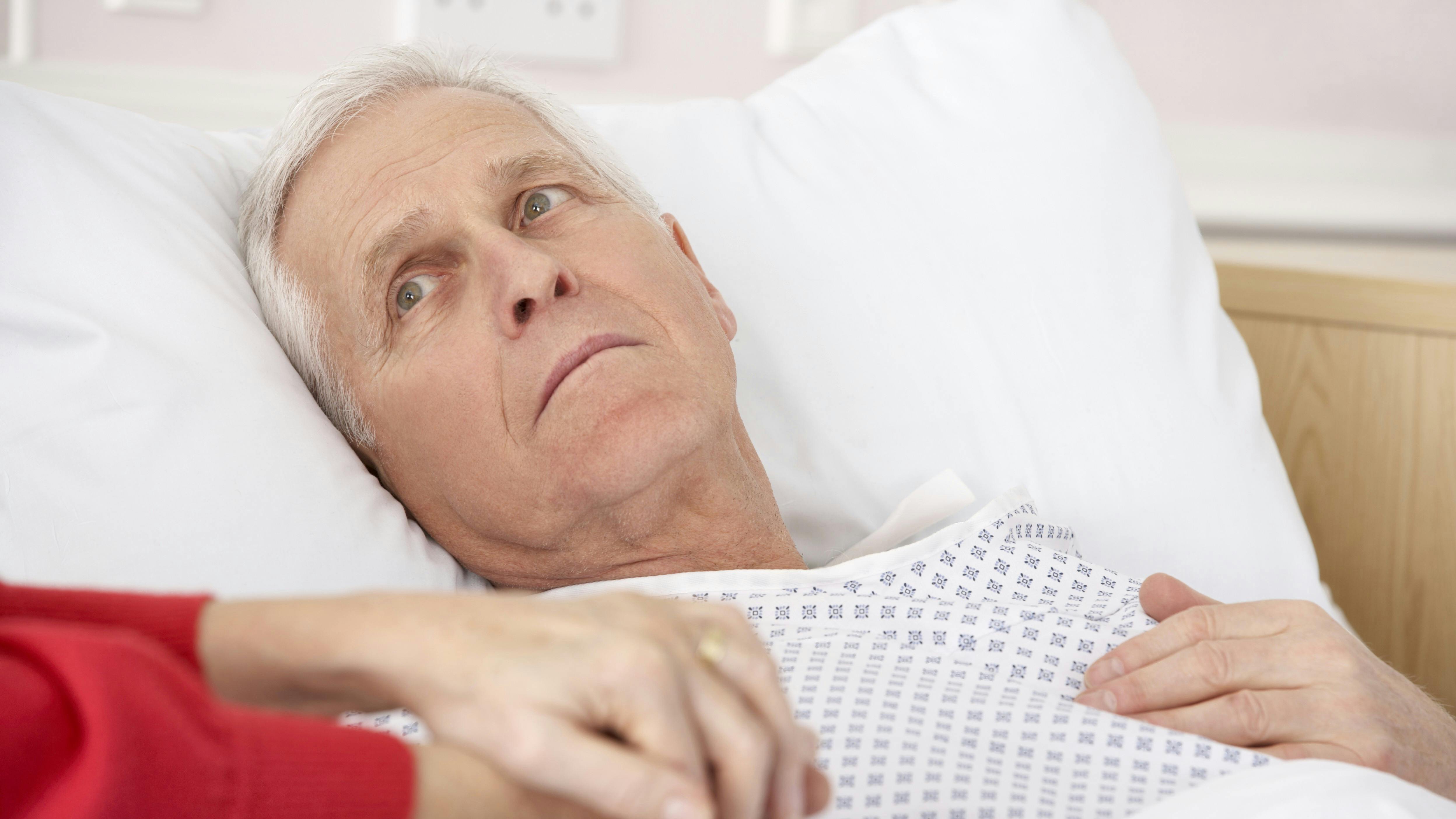 Ældre mand ligger syg i hospitalsseng, mens han holder sin kone i hånden