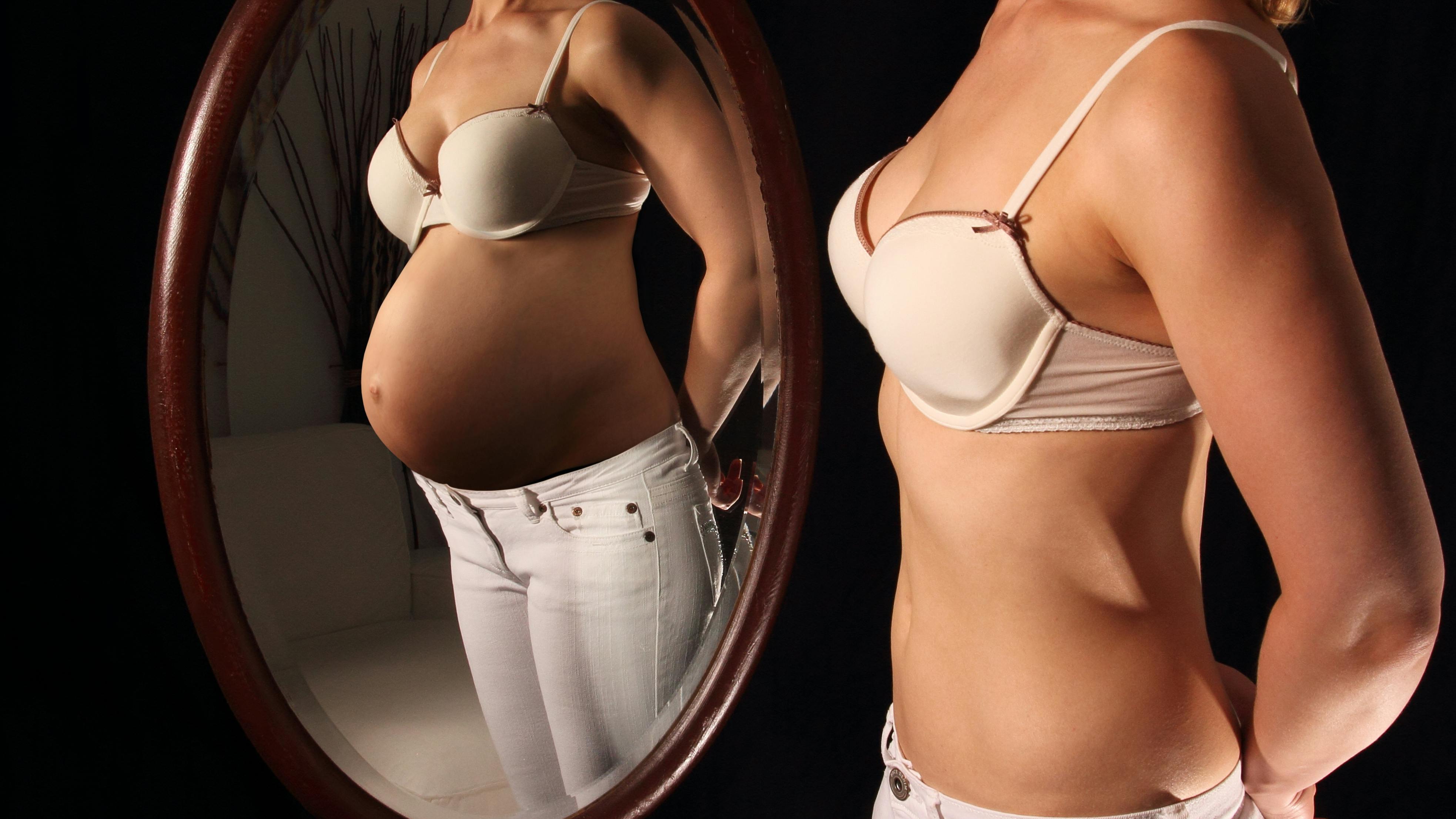 Ung kvinde med flad mave ser på sit spejlbillede, hvor hun er gravid