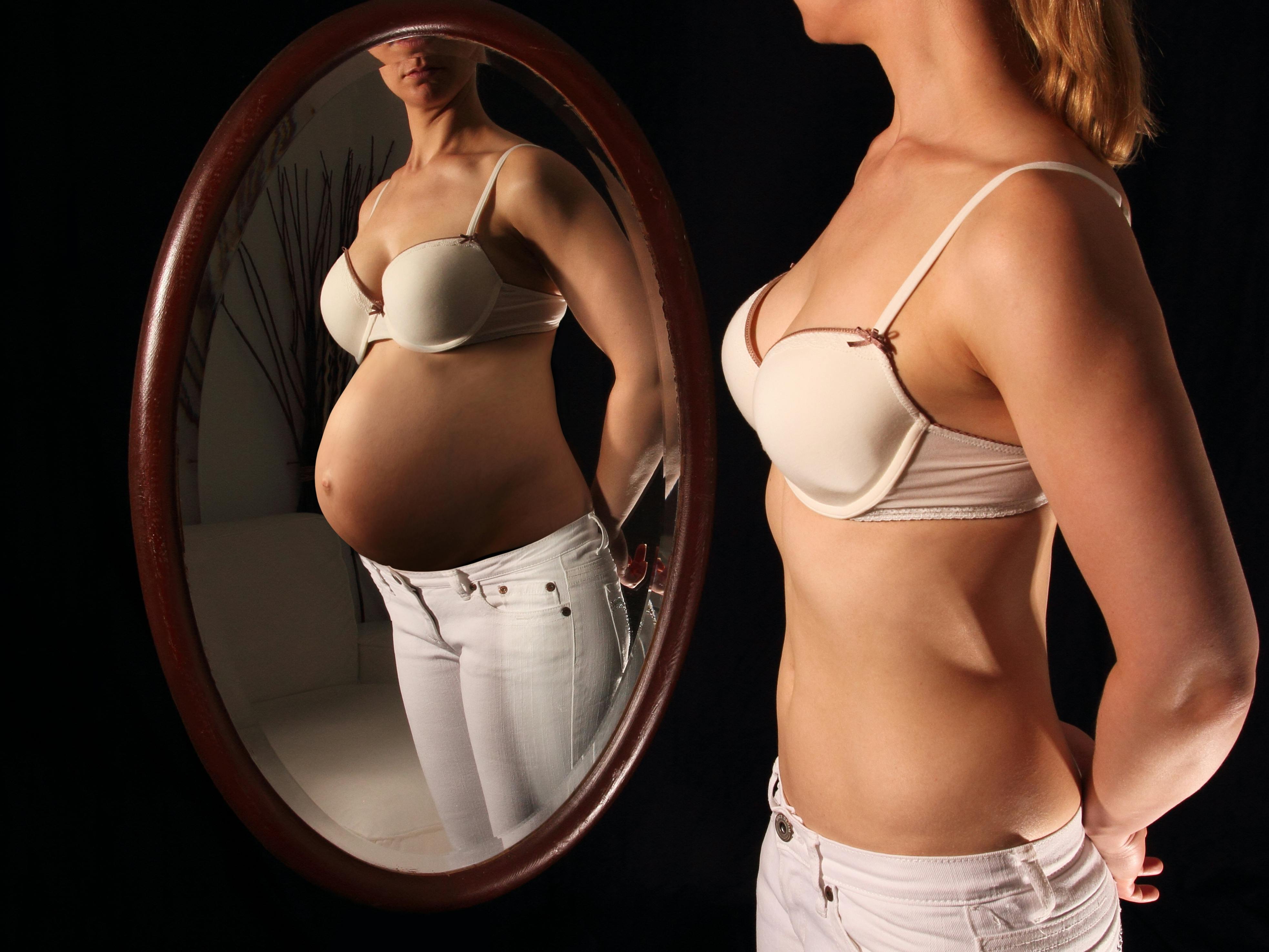 Ung kvinde med flad mave ser på sit spejlbillede, hvor hun er gravid