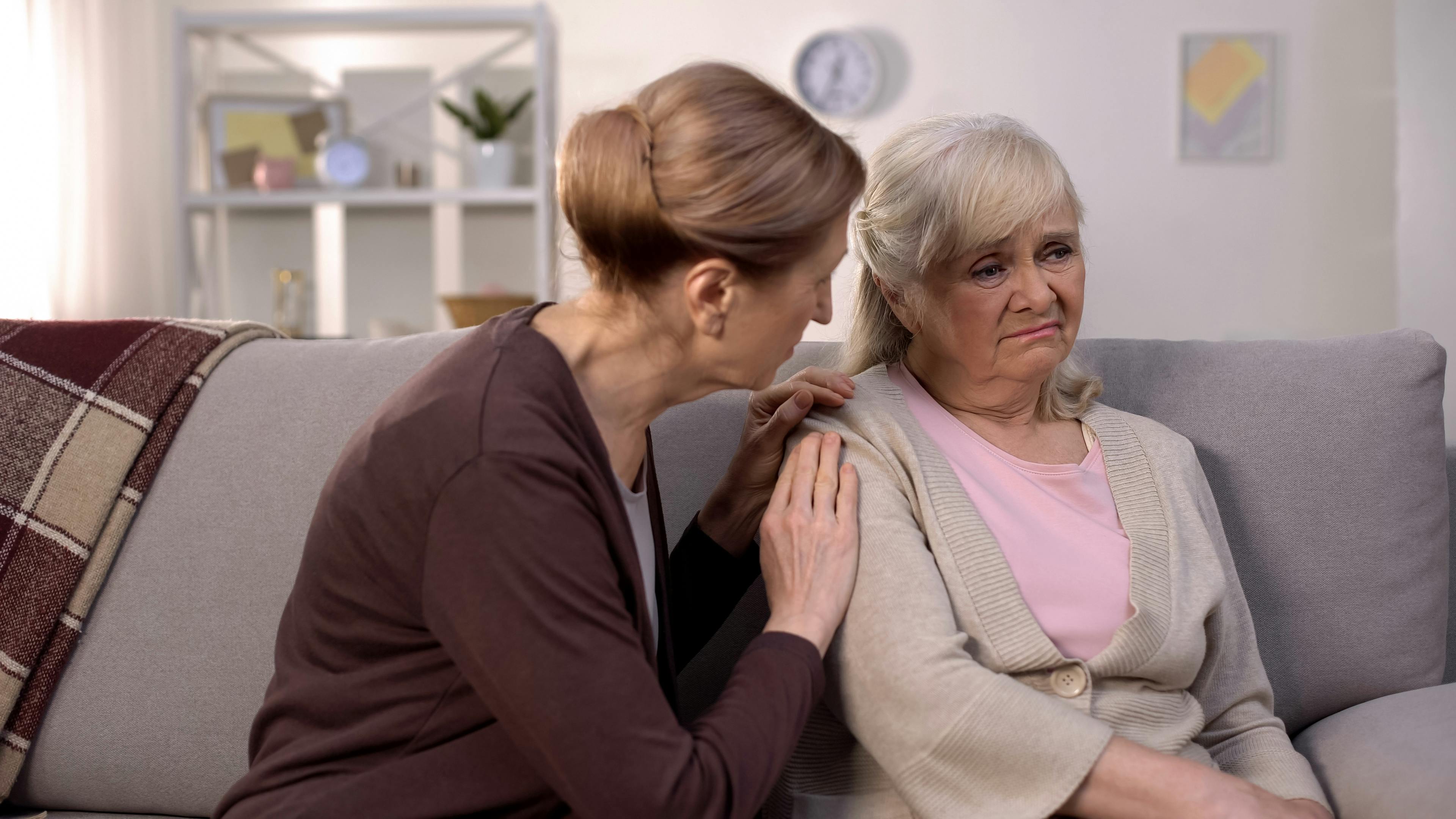 To ældre veninder sidder sammen i en sofa, og den ene holder omsorgsfuldt den anden på skulderen.