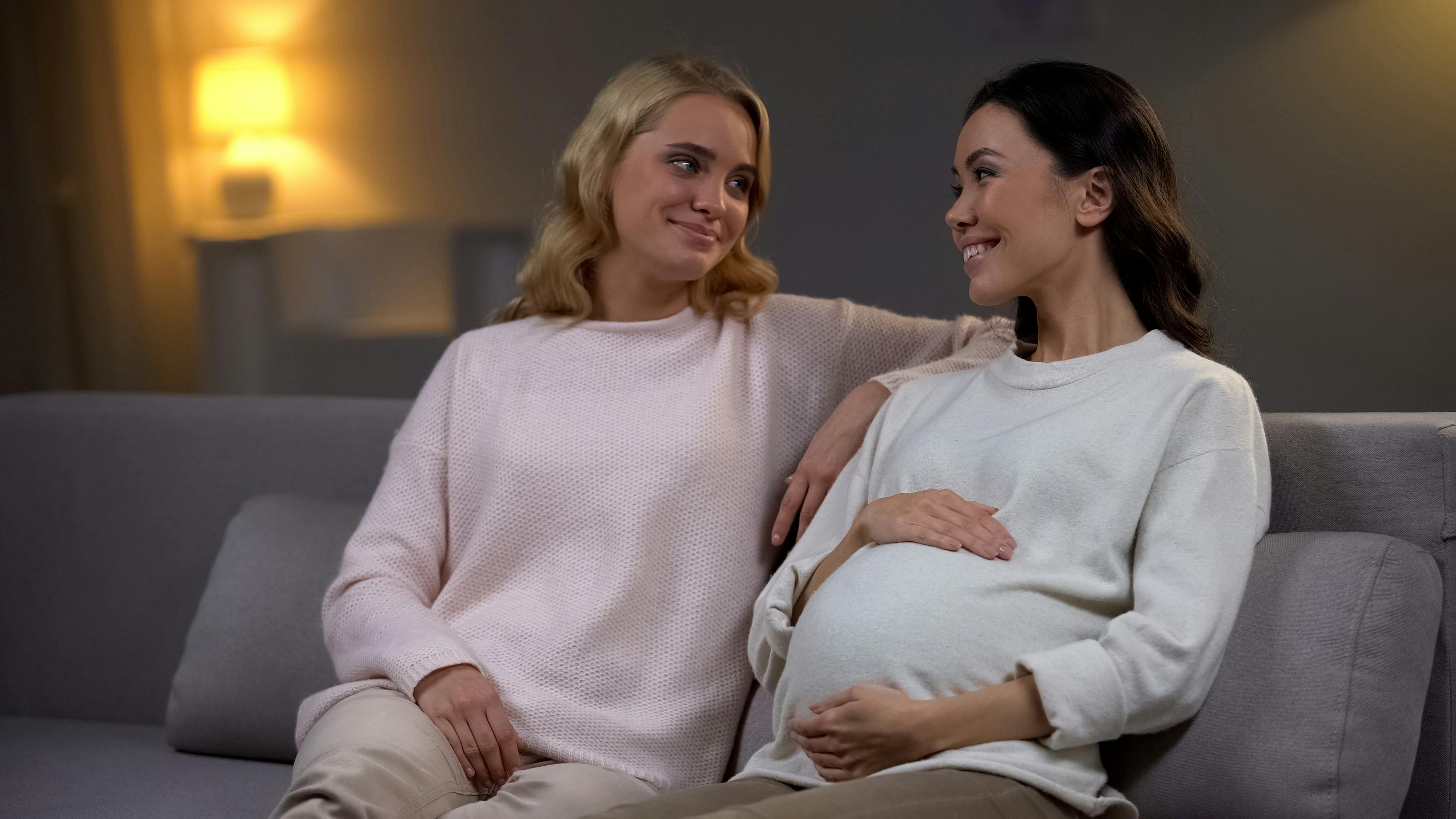 To veninder sidder sammen i en sofa. Den ene er gravid og ser glad ud. 