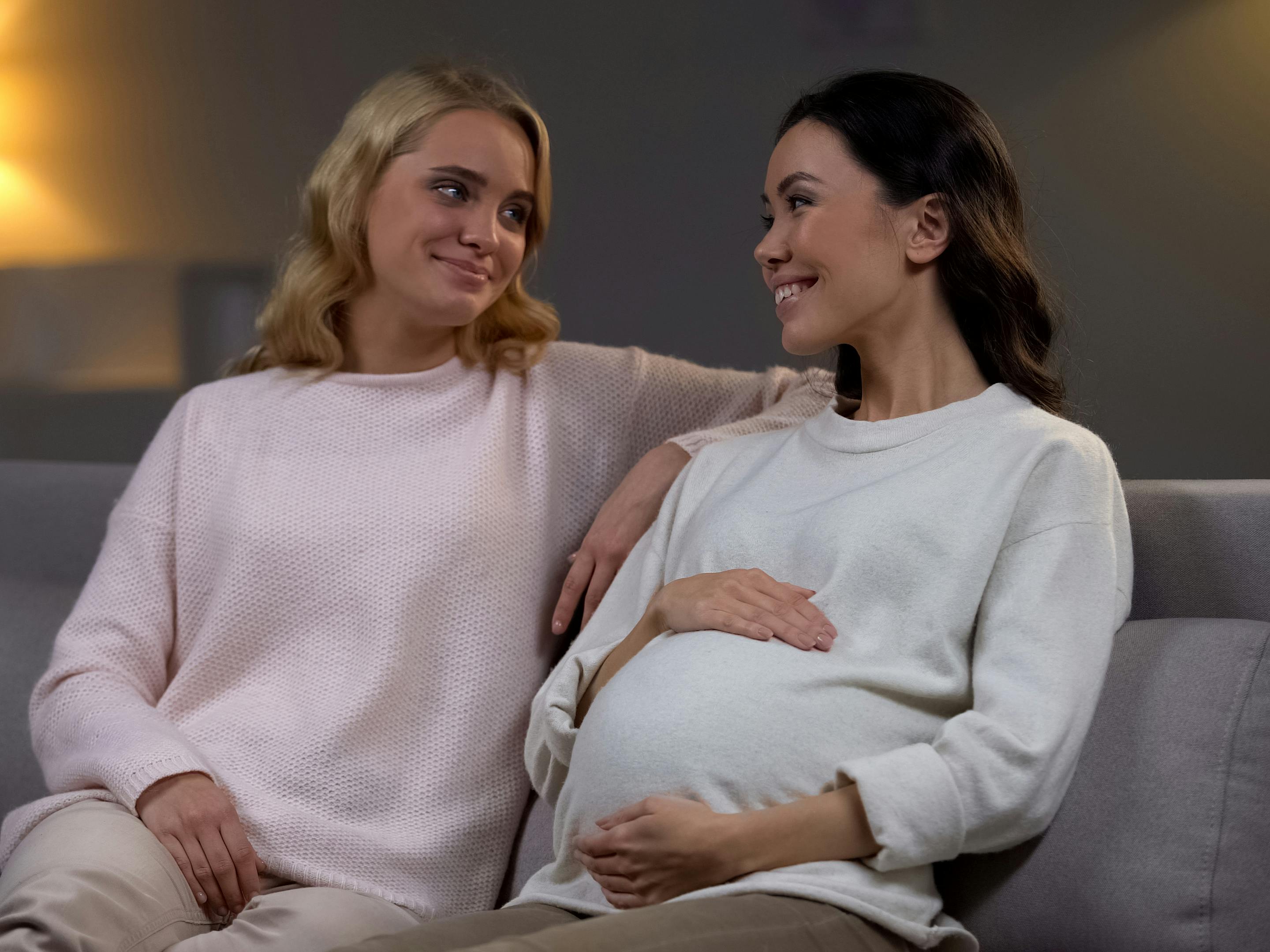 To veninder sidder sammen i en sofa. Den ene er gravid og ser glad ud. 