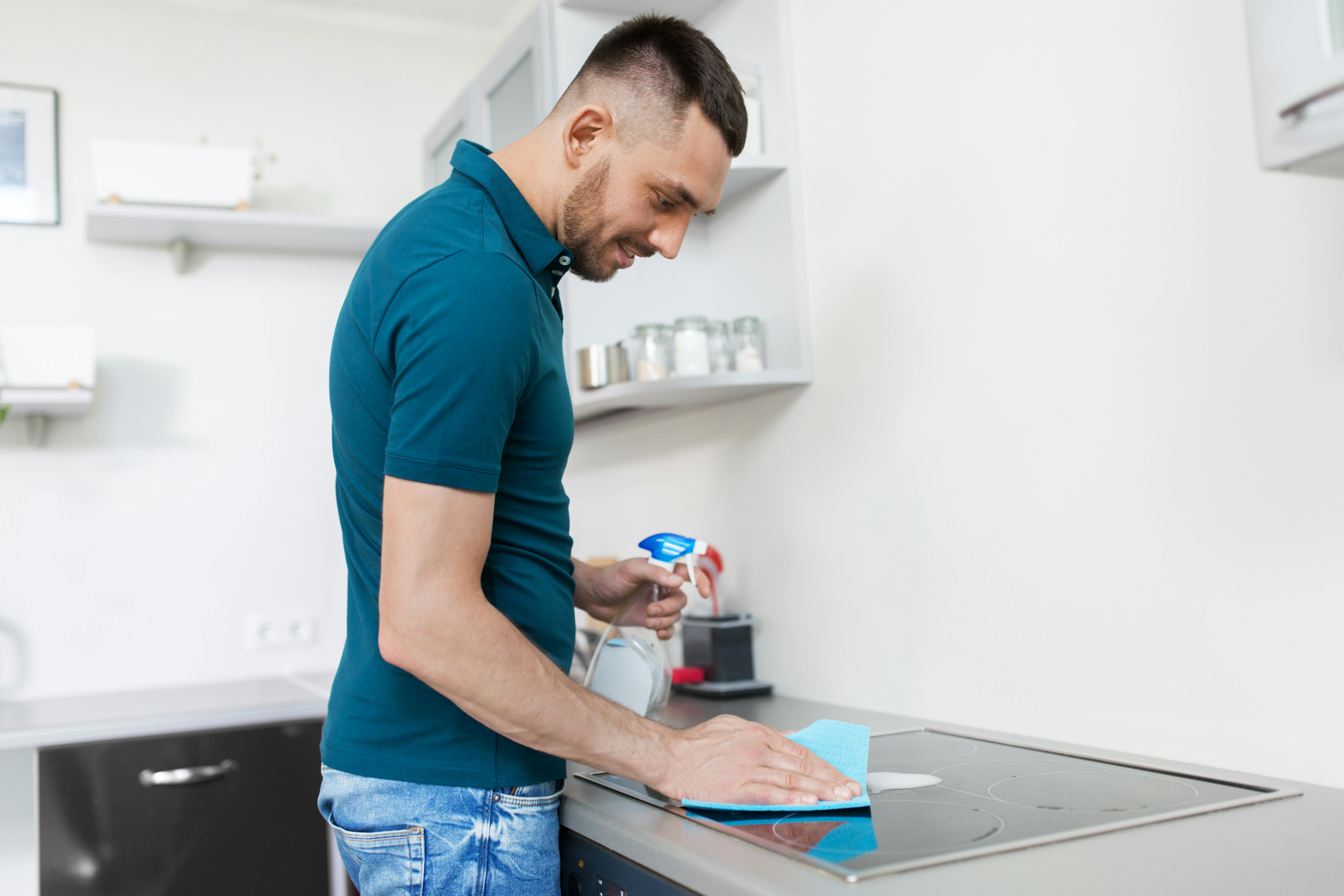 cleaning, household and people concept - man with spray cleaner and cloth wiping cooker at home kitchen