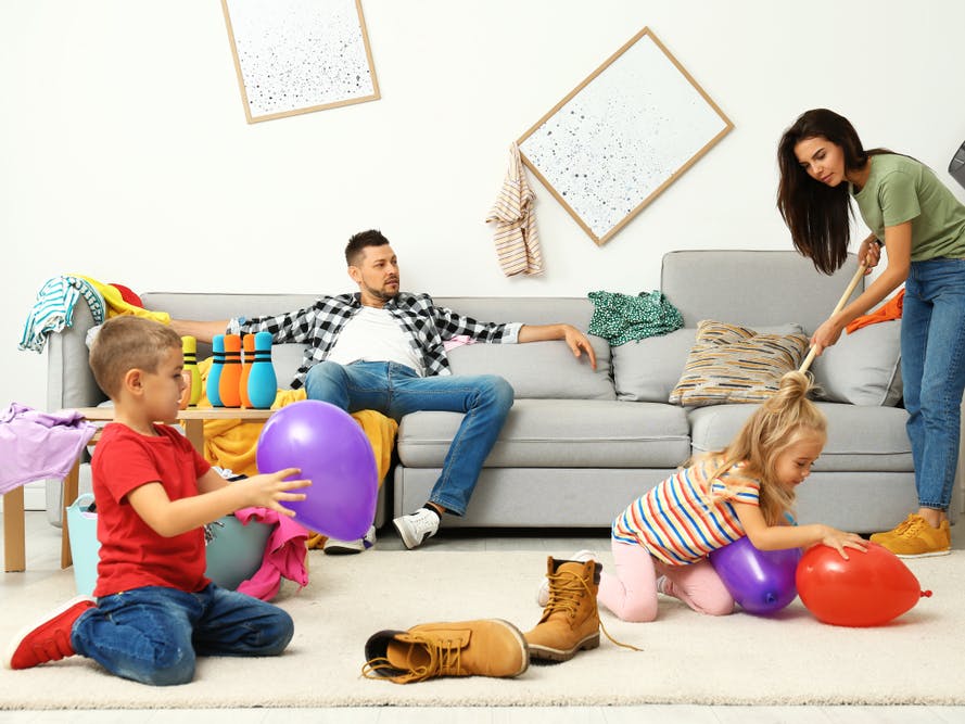 Kvinde støvsuger sit rodede hjem, mens hendes mand sidder i sofaen, og børnene leger på gulvet.