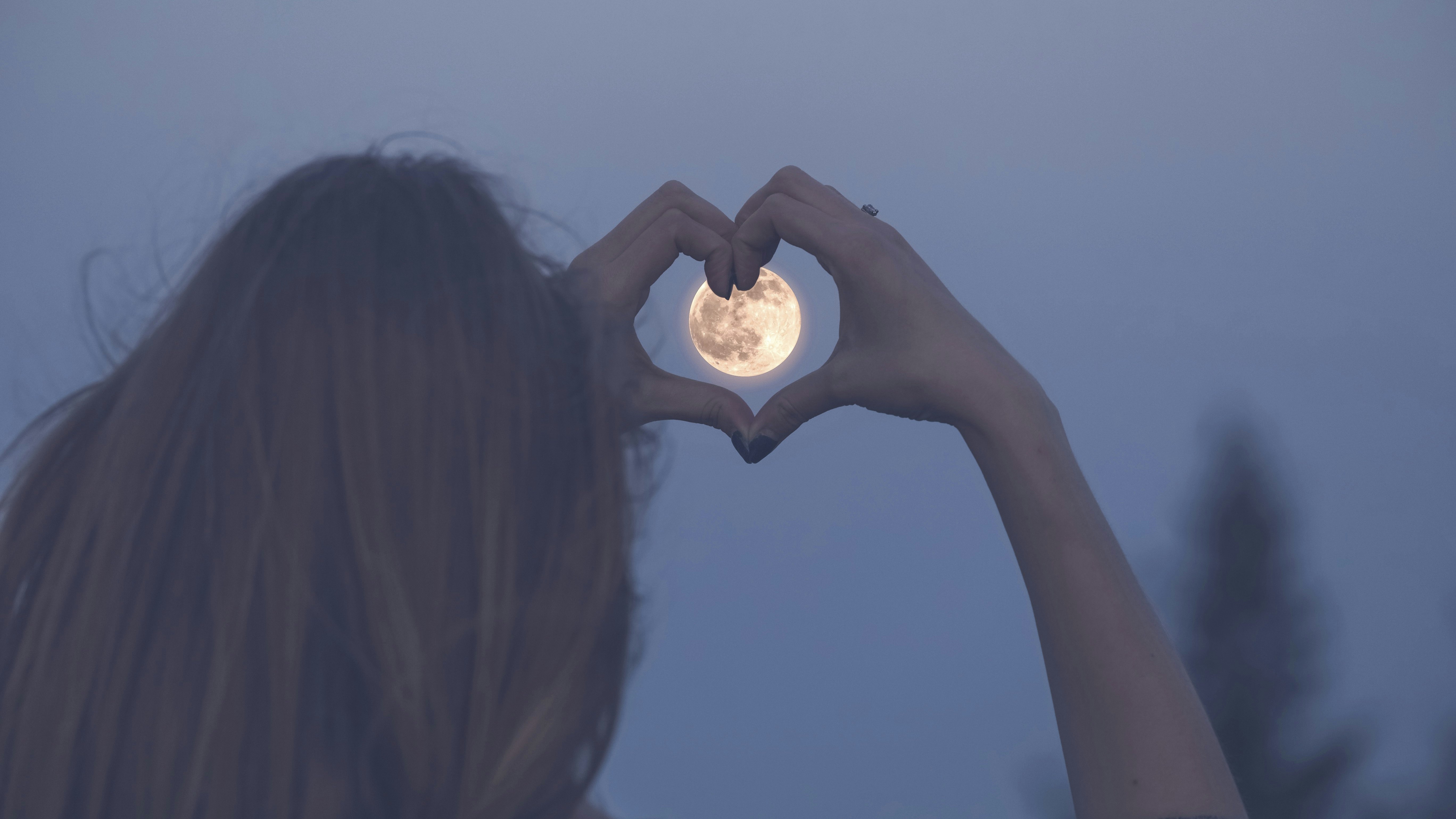 Kvinde former et hjerte med sine fingre foran en fuldmåne