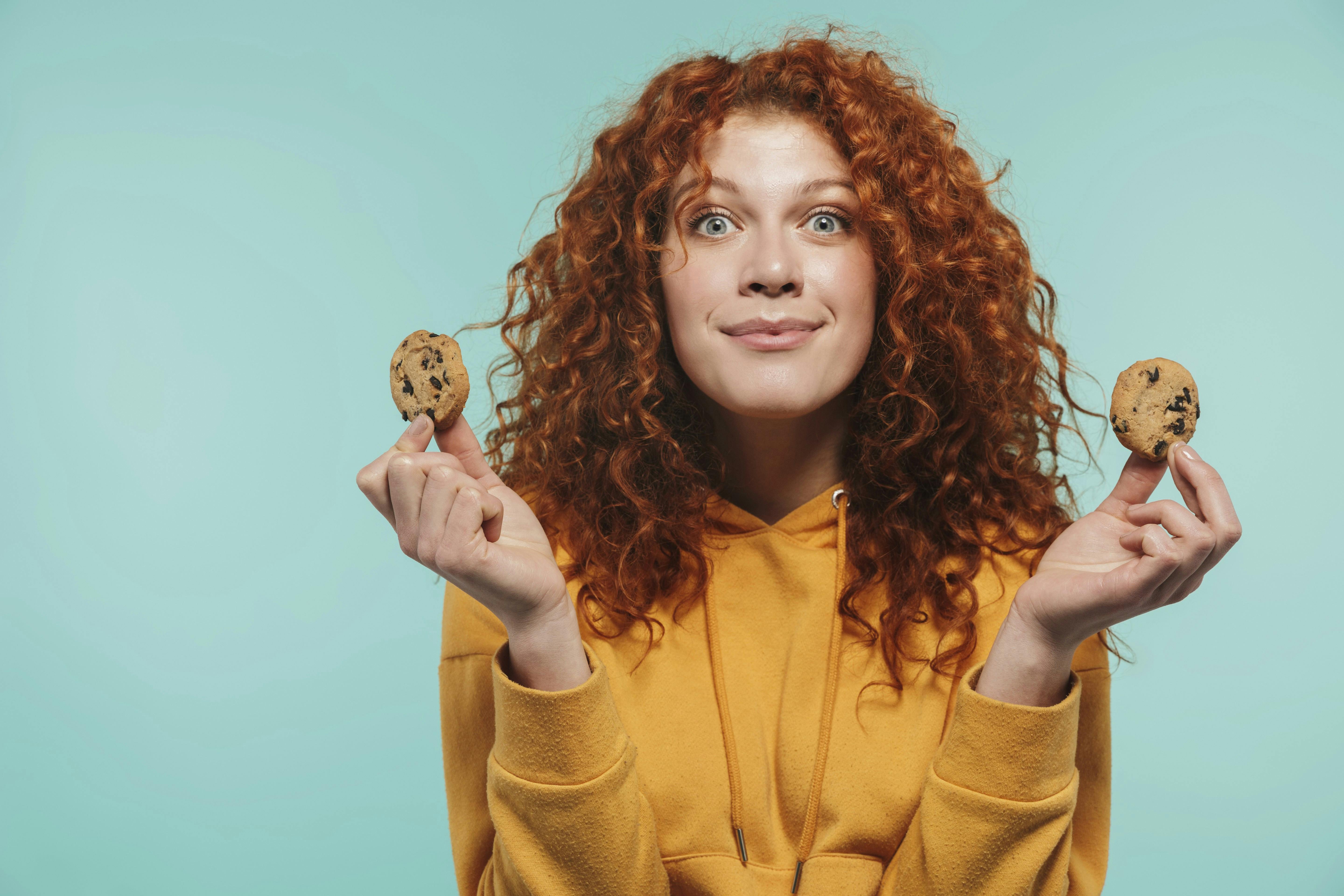 Portrait of happy redhead woman 20s smiling and eating sweet cookies isolated over blue background