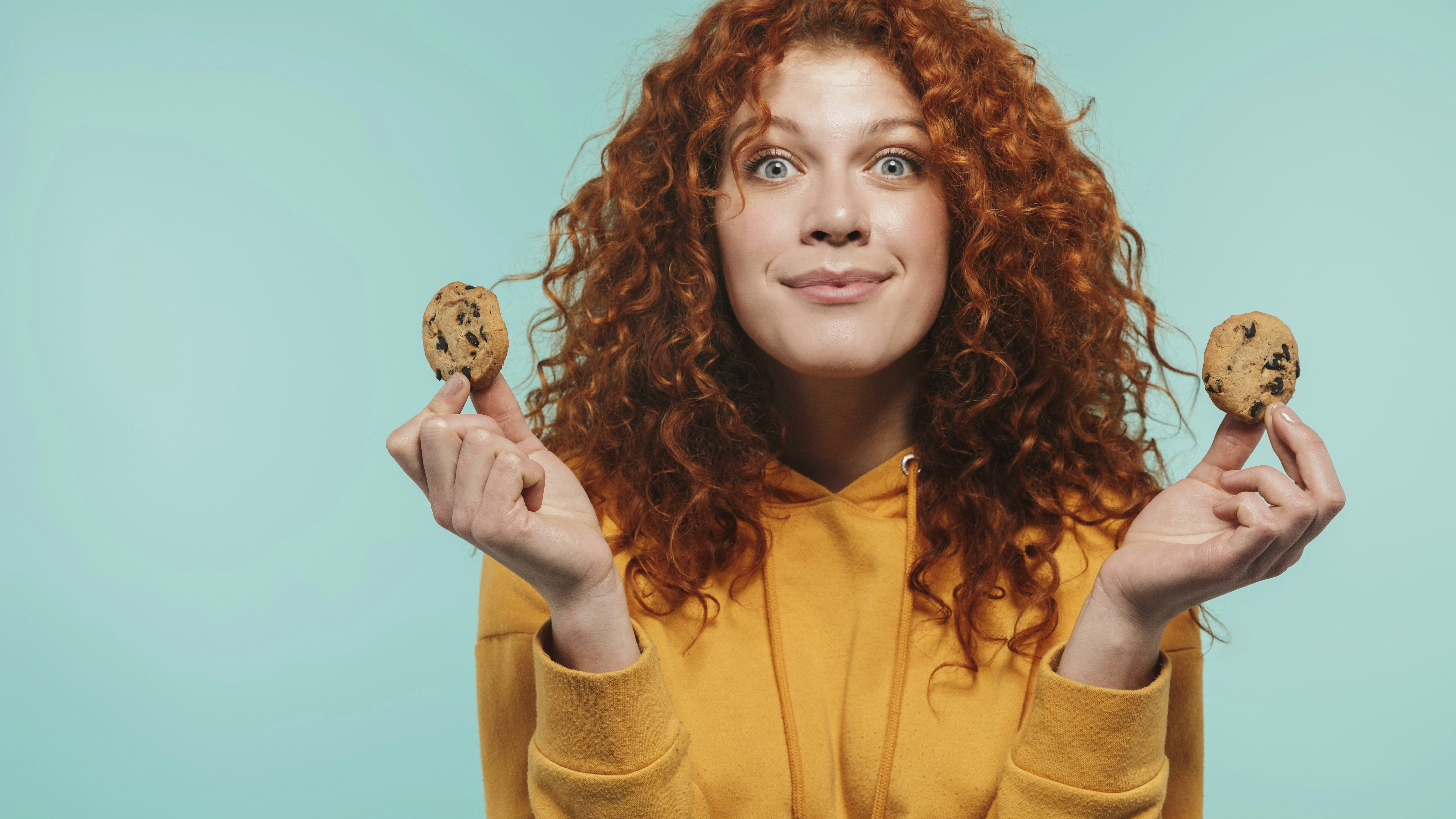Portrait of happy redhead woman 20s smiling and eating sweet cookies isolated over blue background