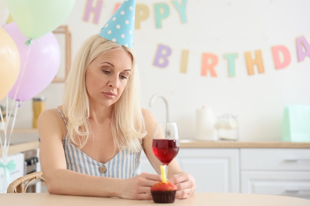 Midaldrende kvinde sidder med vin og fødselshat og ser trist ud.