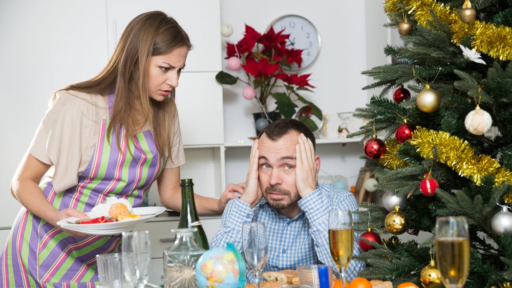 Kvinde ser vredt på sin mand, der sidder vel julebordet med et juletræ i baggrunden.