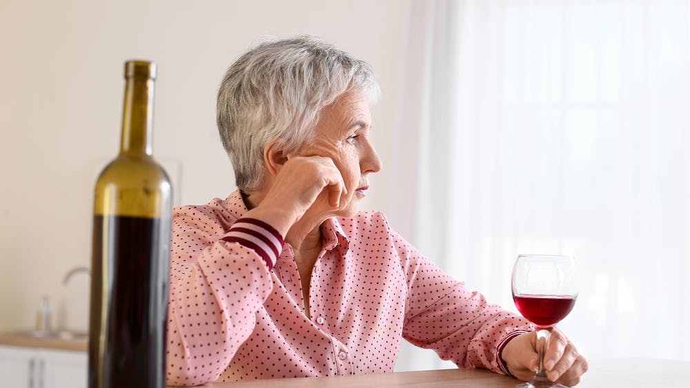 Ældre kvinde sidder med et glas og en flaske vin og ser trist ud.