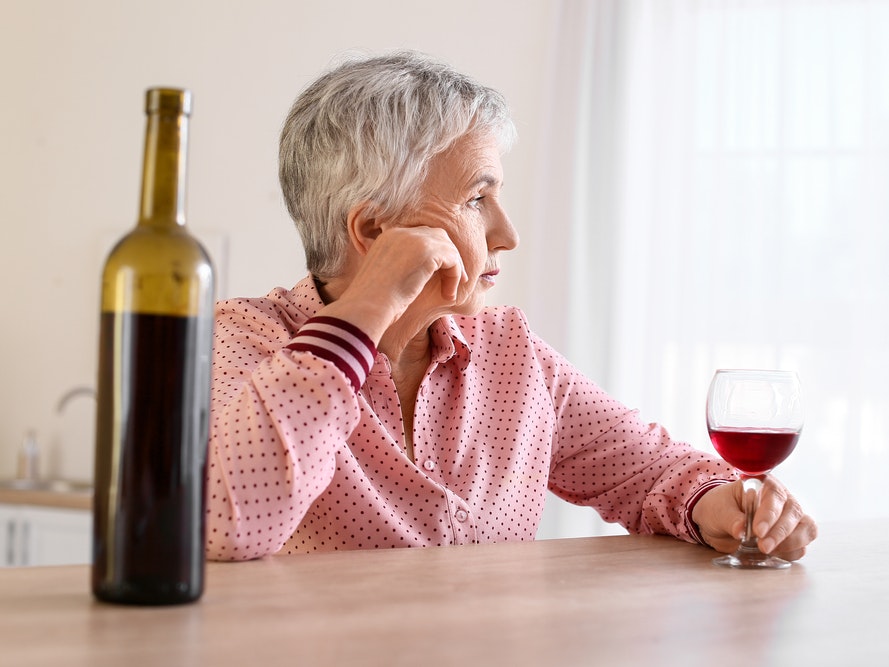 Ældre kvinde sidder med et glas og en flaske vin og ser trist ud.