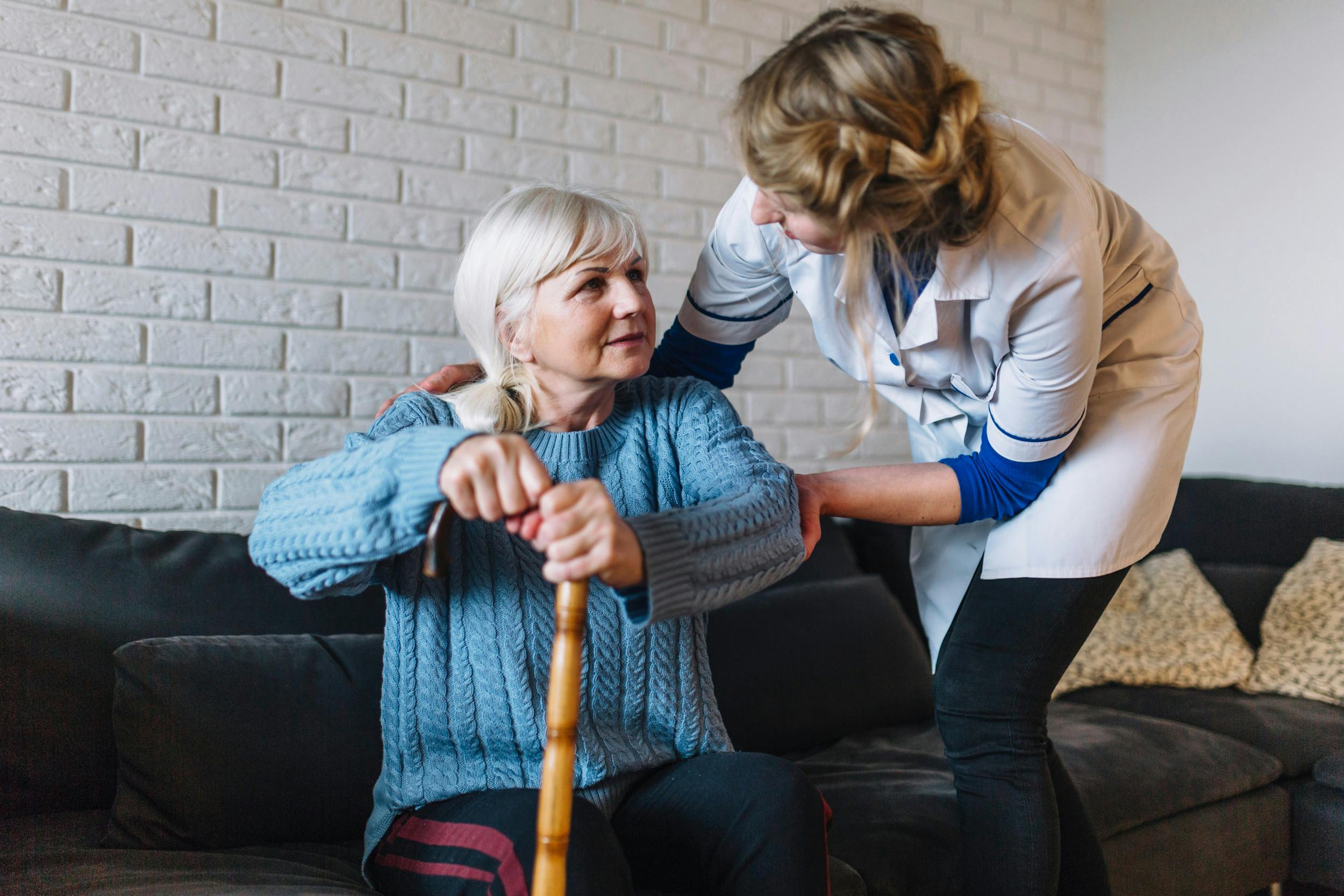 Plejehjemsansat hjælper ældre kvinde med at rejse sig fra sofaen.