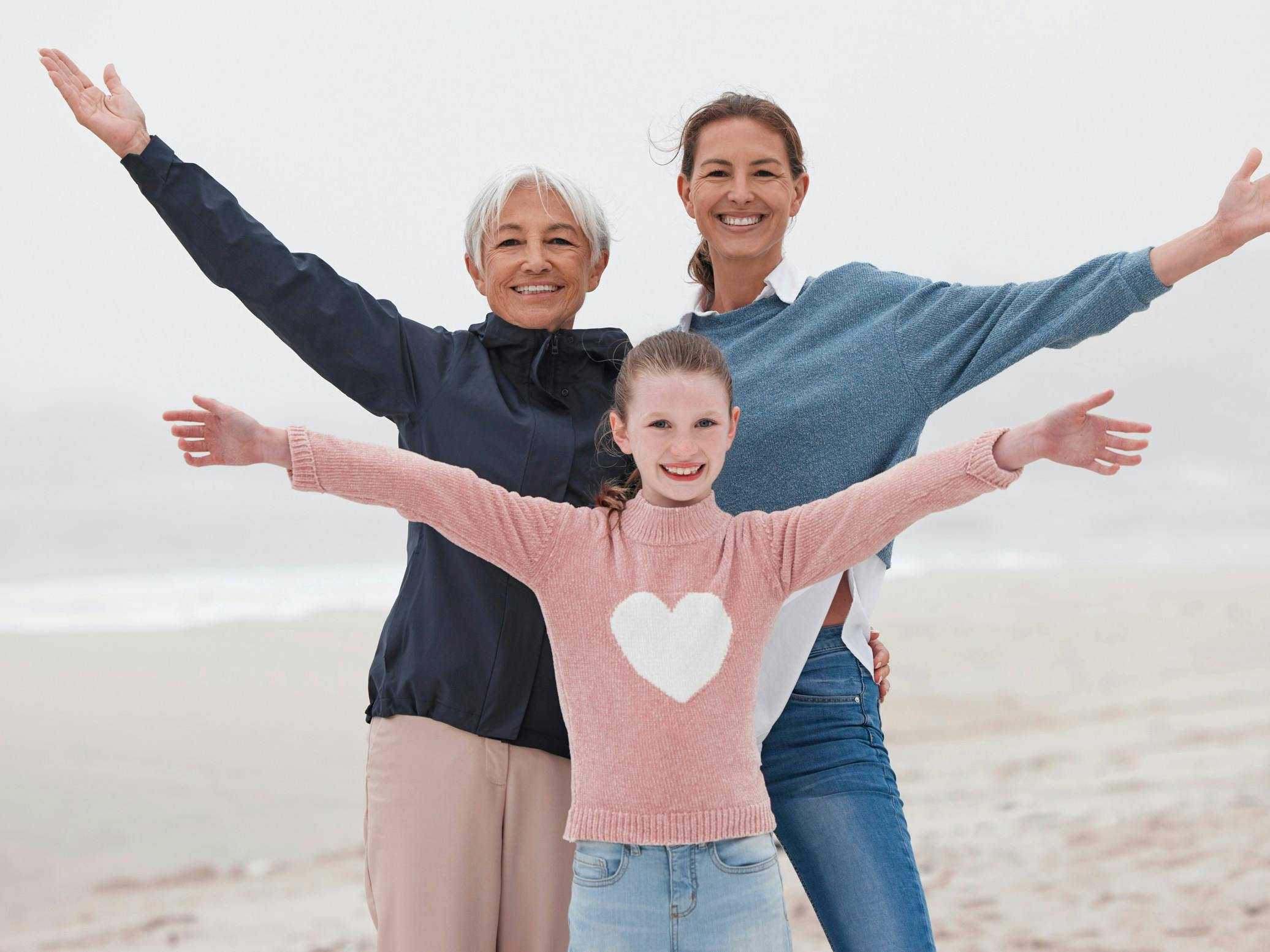 Mormor, mor og datter står på stranden med udbredte arme og store smil.