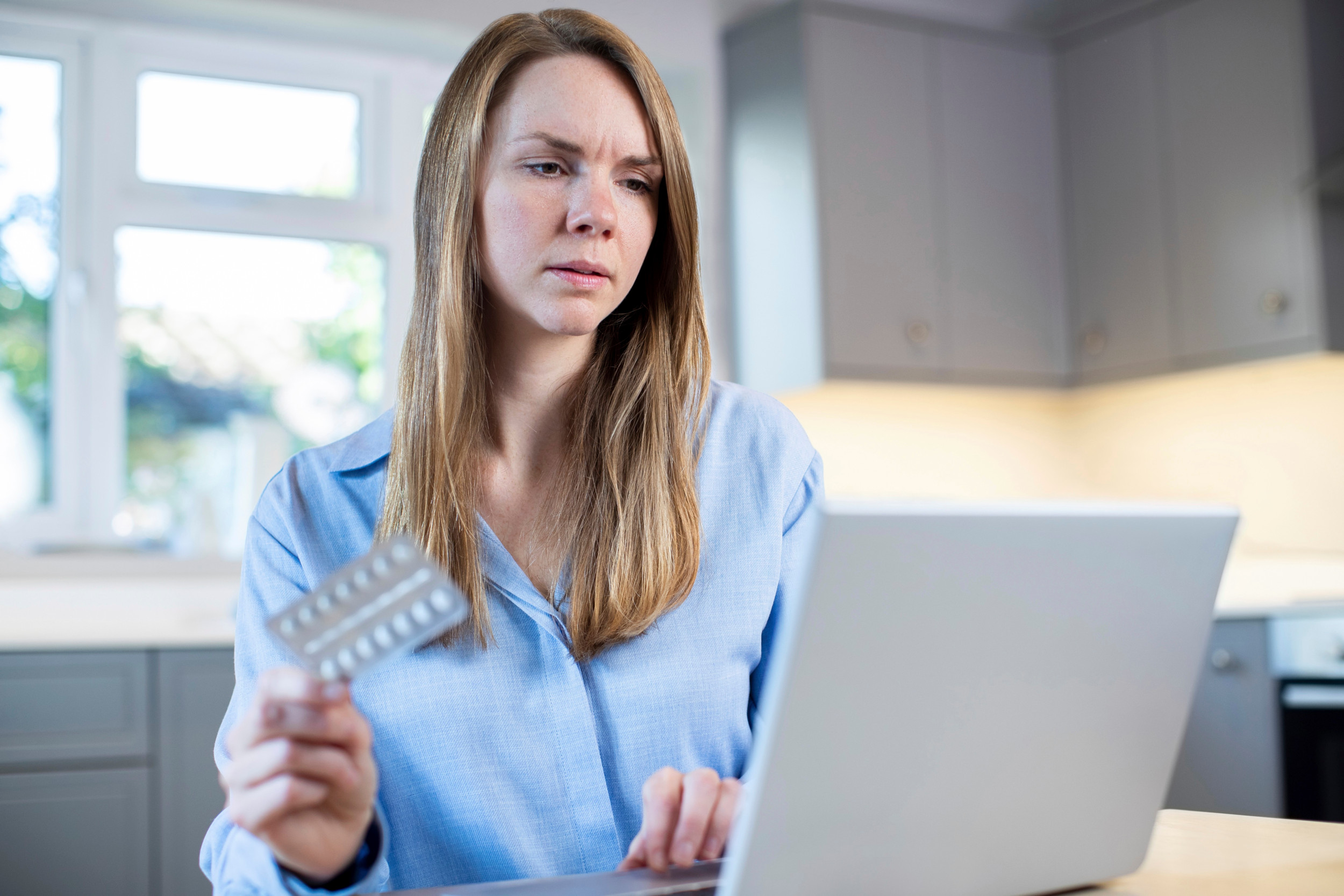 Kvinde sidder foran sin bærbare computer og holder piller i hånden.