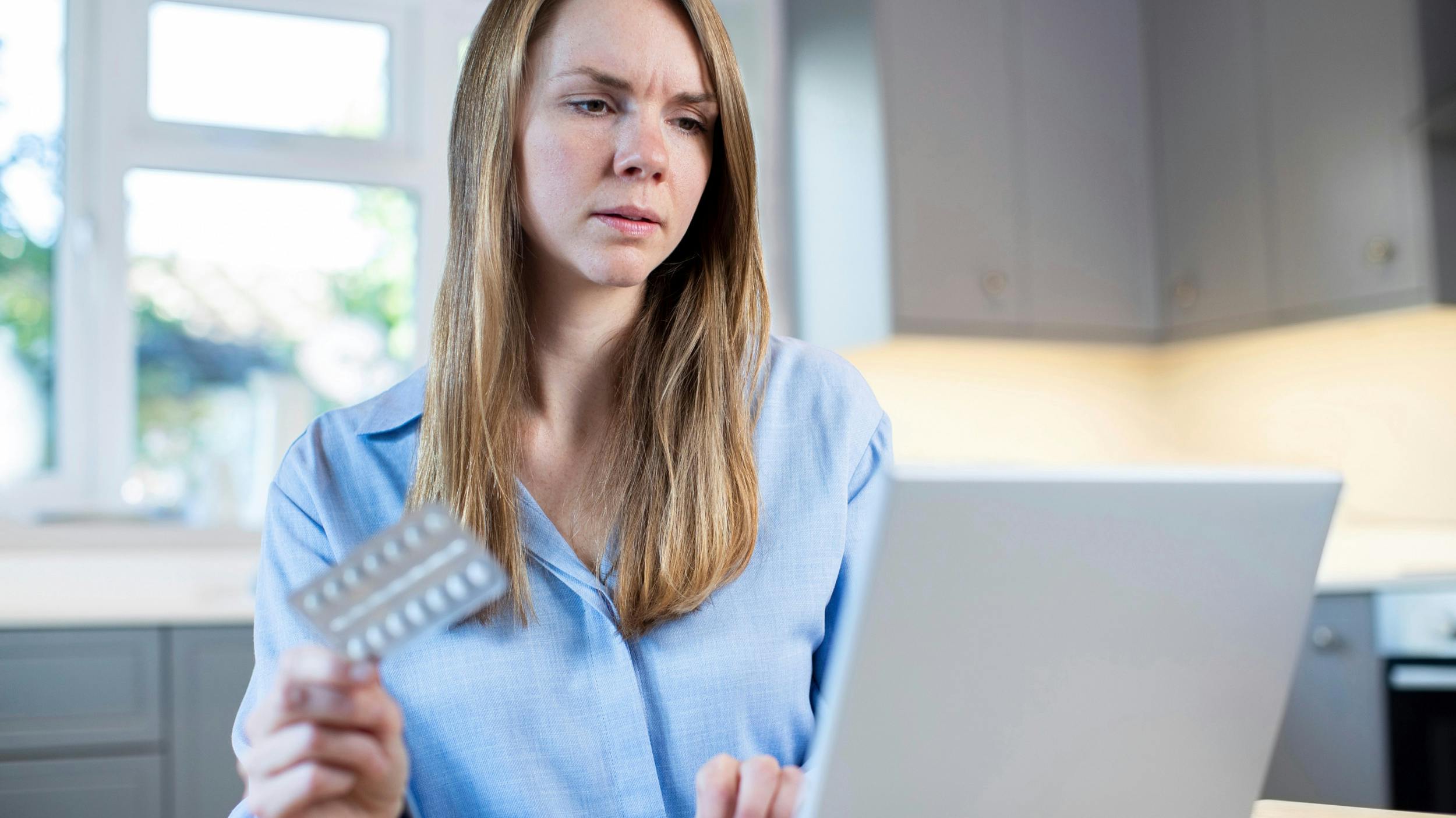 Kvinde sidder foran sin bærbare computer og holder piller i hånde