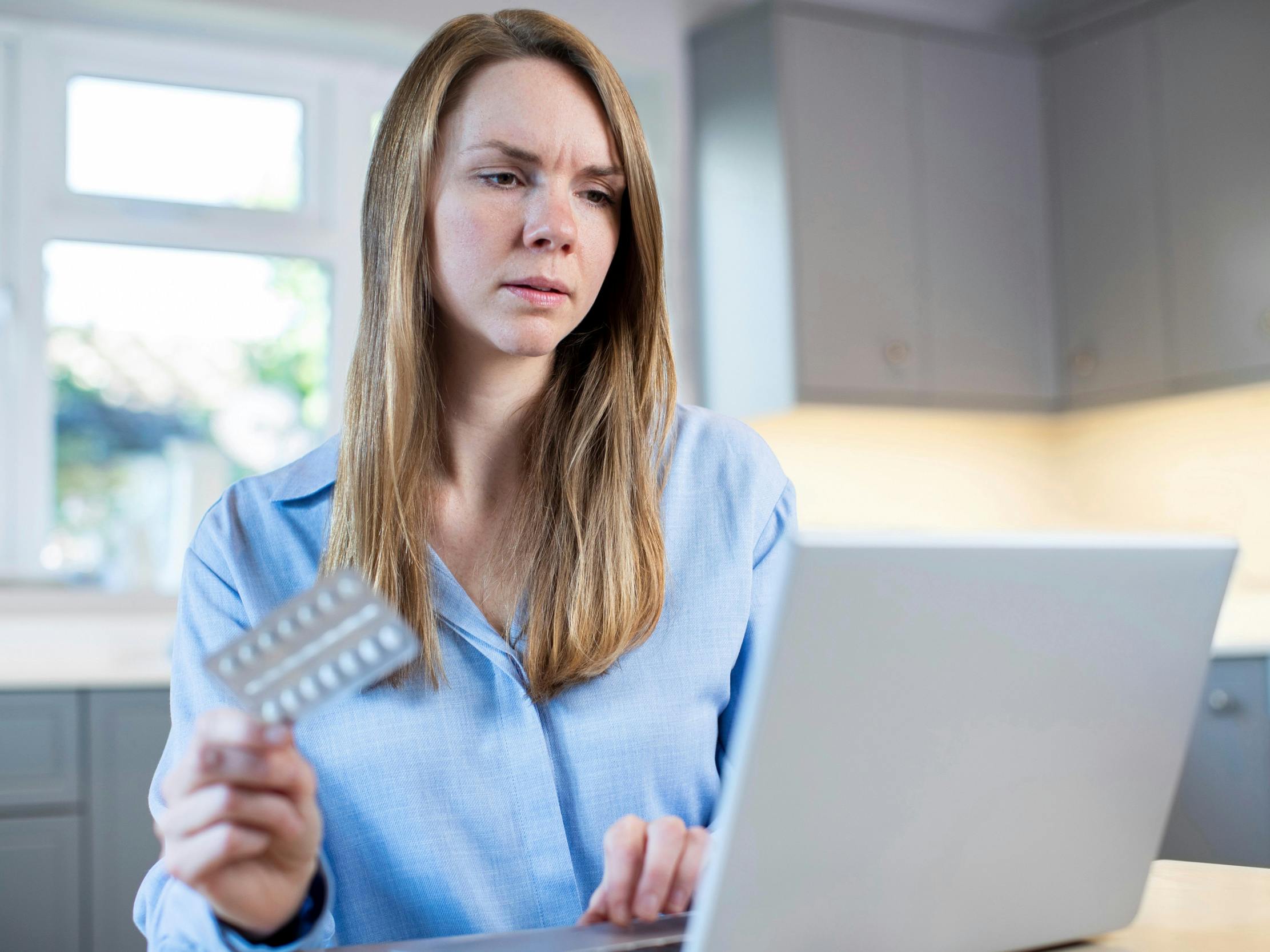Kvinde sidder foran sin bærbare computer og holder piller i hånden.