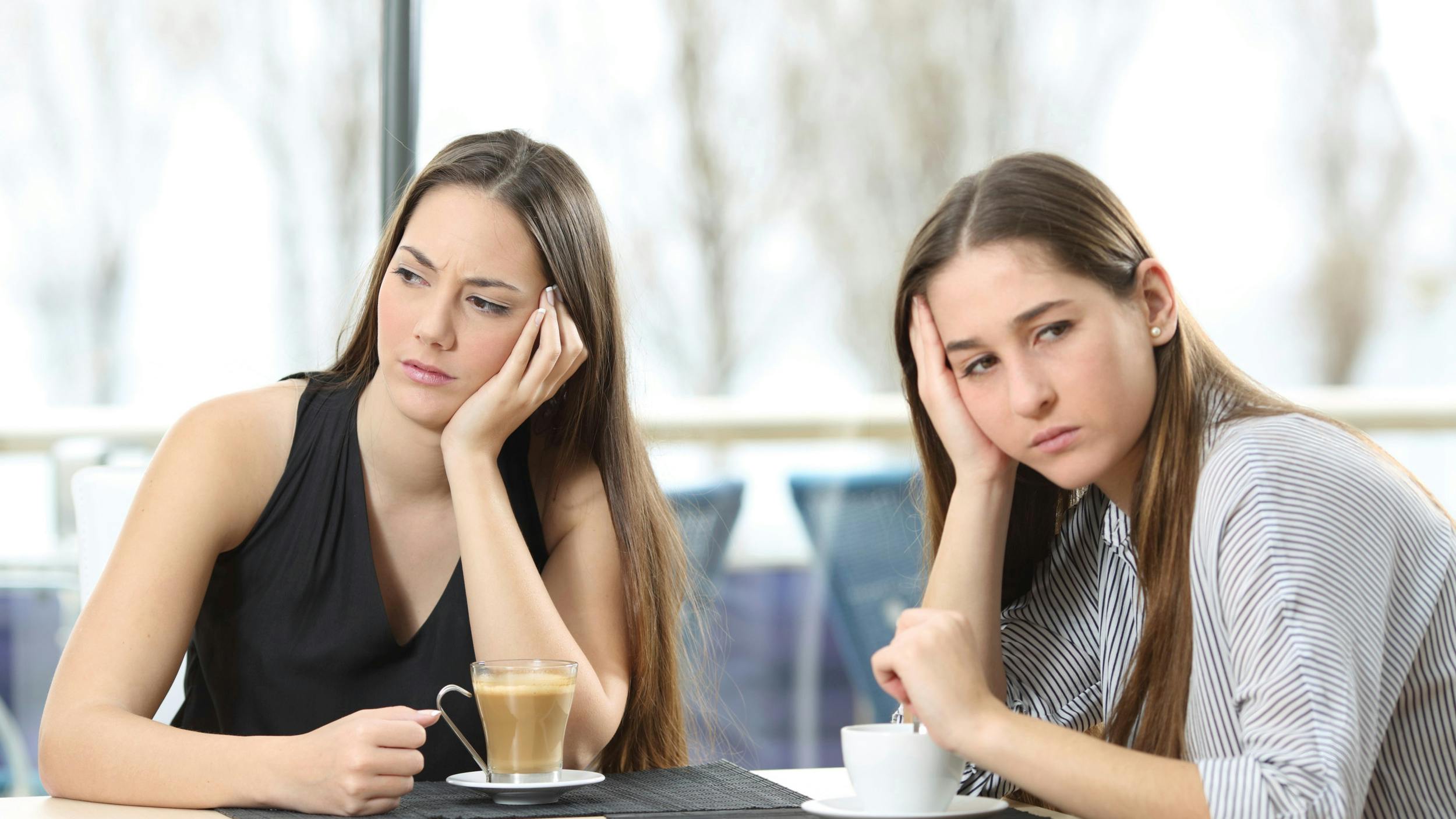 To kvinder sidder med hver sin kop kaffe, mens de ser hver sin vej med et trist udtryk.