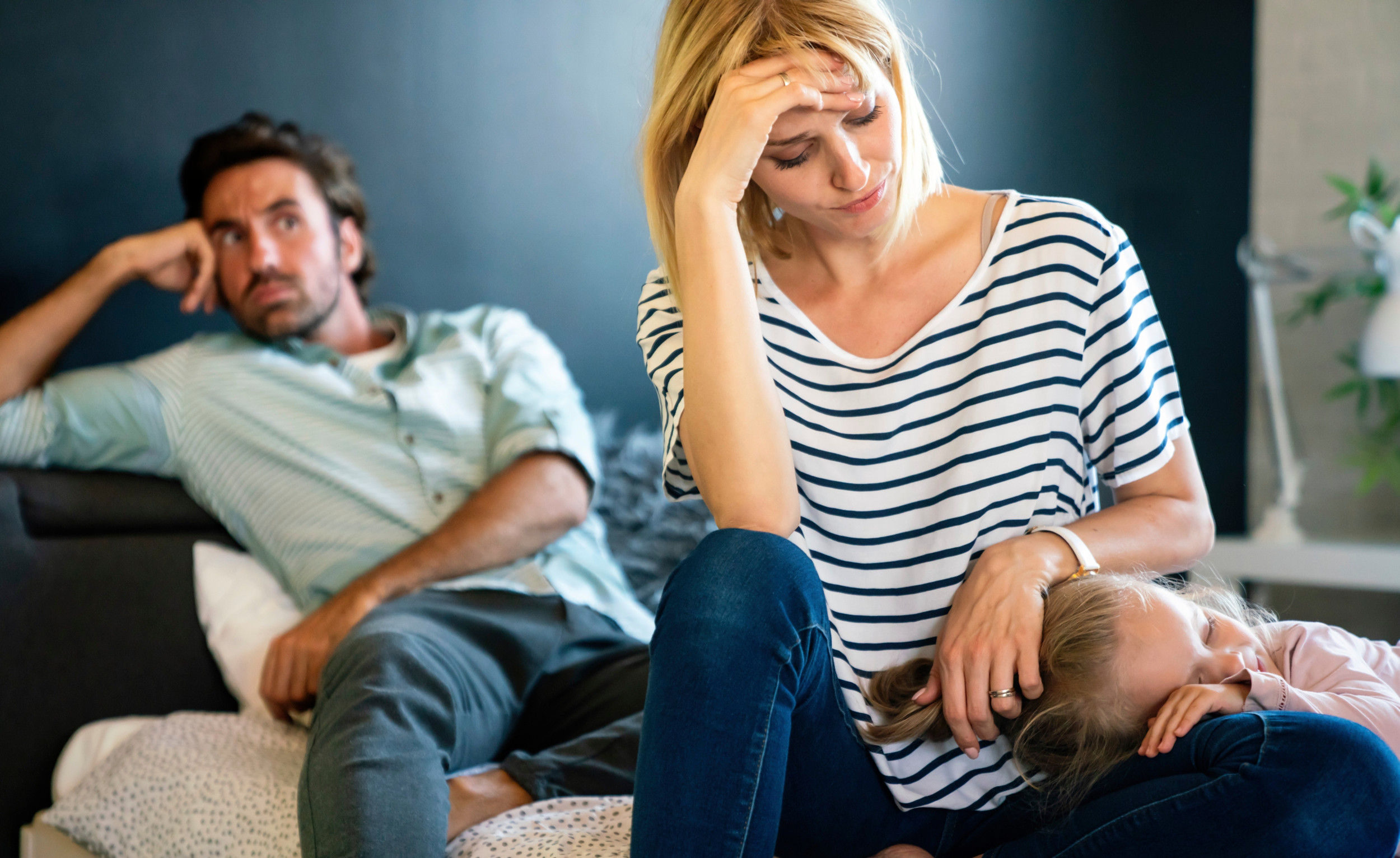Kvinde med datter liggende i skødet ser opgivende ud, mens hendes mand ser til i baggrunden.