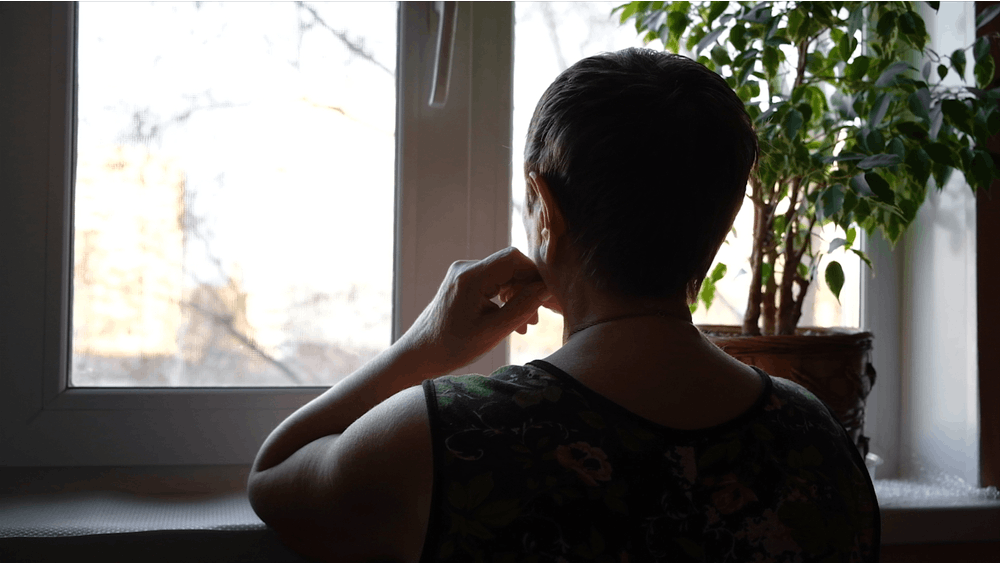 Midaldrende kvinde sidder i tusmørke og ser ud af sit vindue.