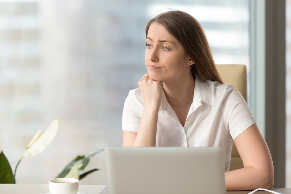 Kvinde sidder ved computeren på sin arbejdsplads og ser eftertænksom ud i luften.