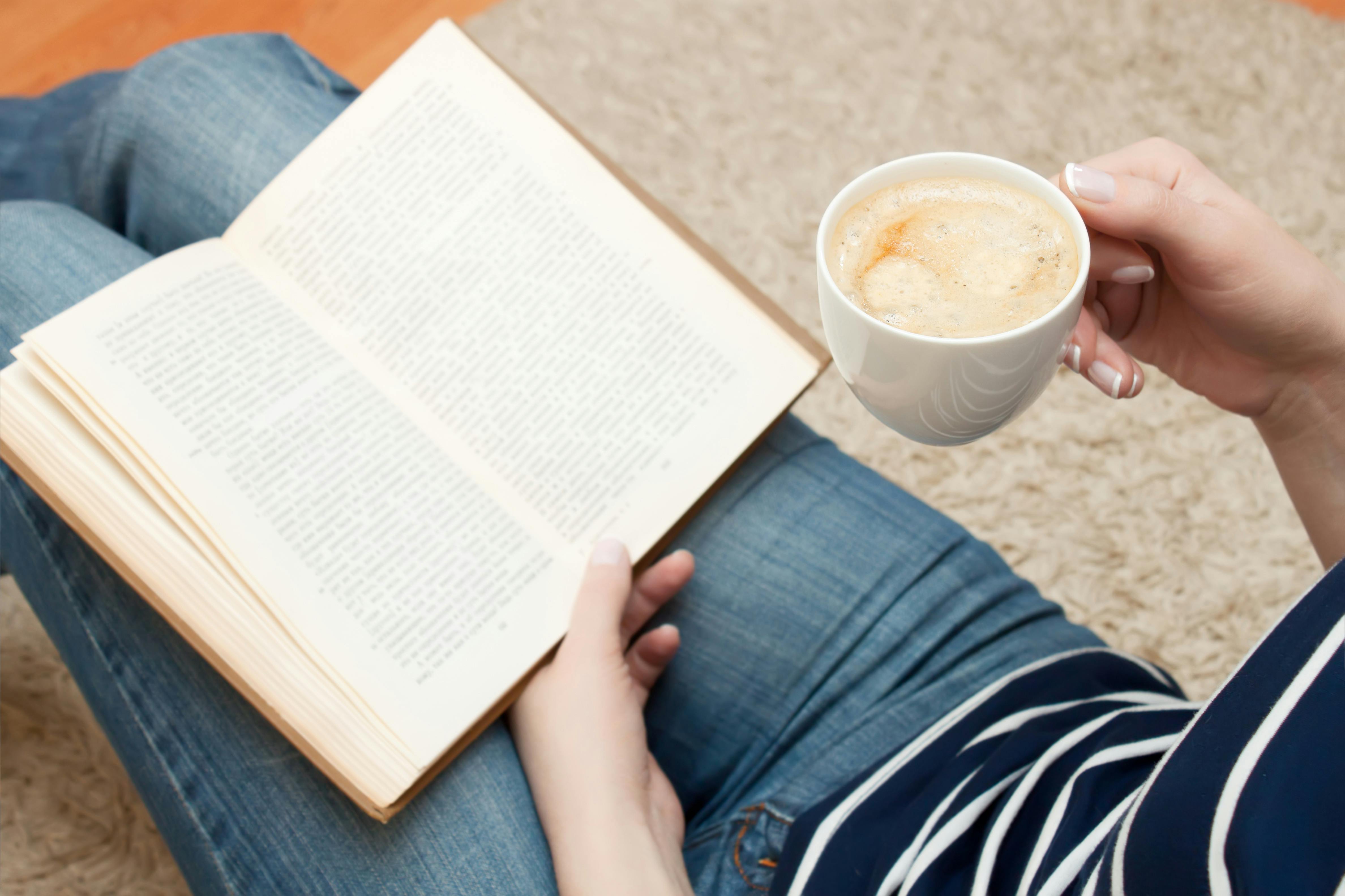 Kvinden læser en bog, mens hun sidder på gulvet og holder en kop kaffe