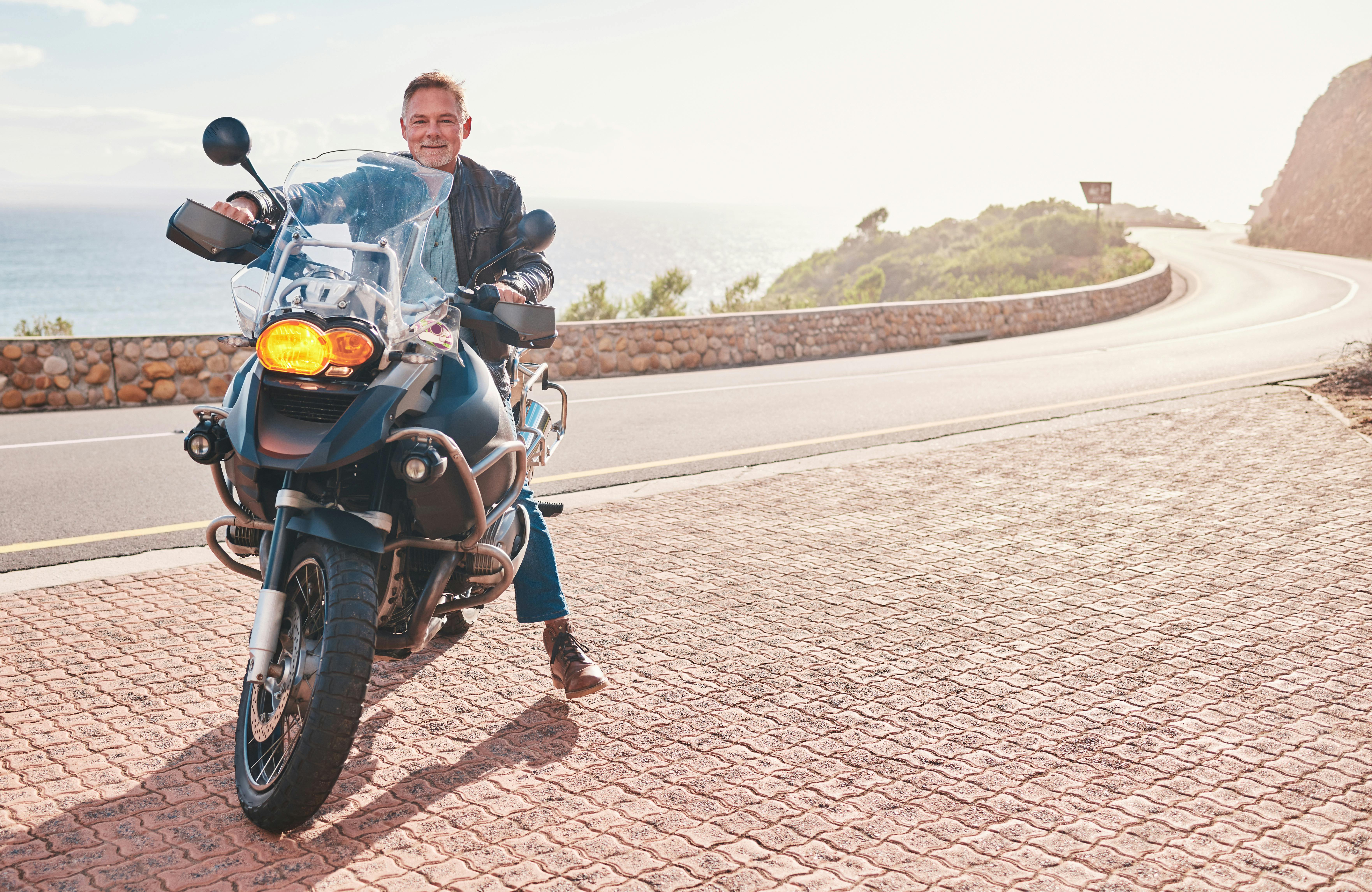 Midaldrende mand holder på sin motorcykel på en bjergvej langs havet.