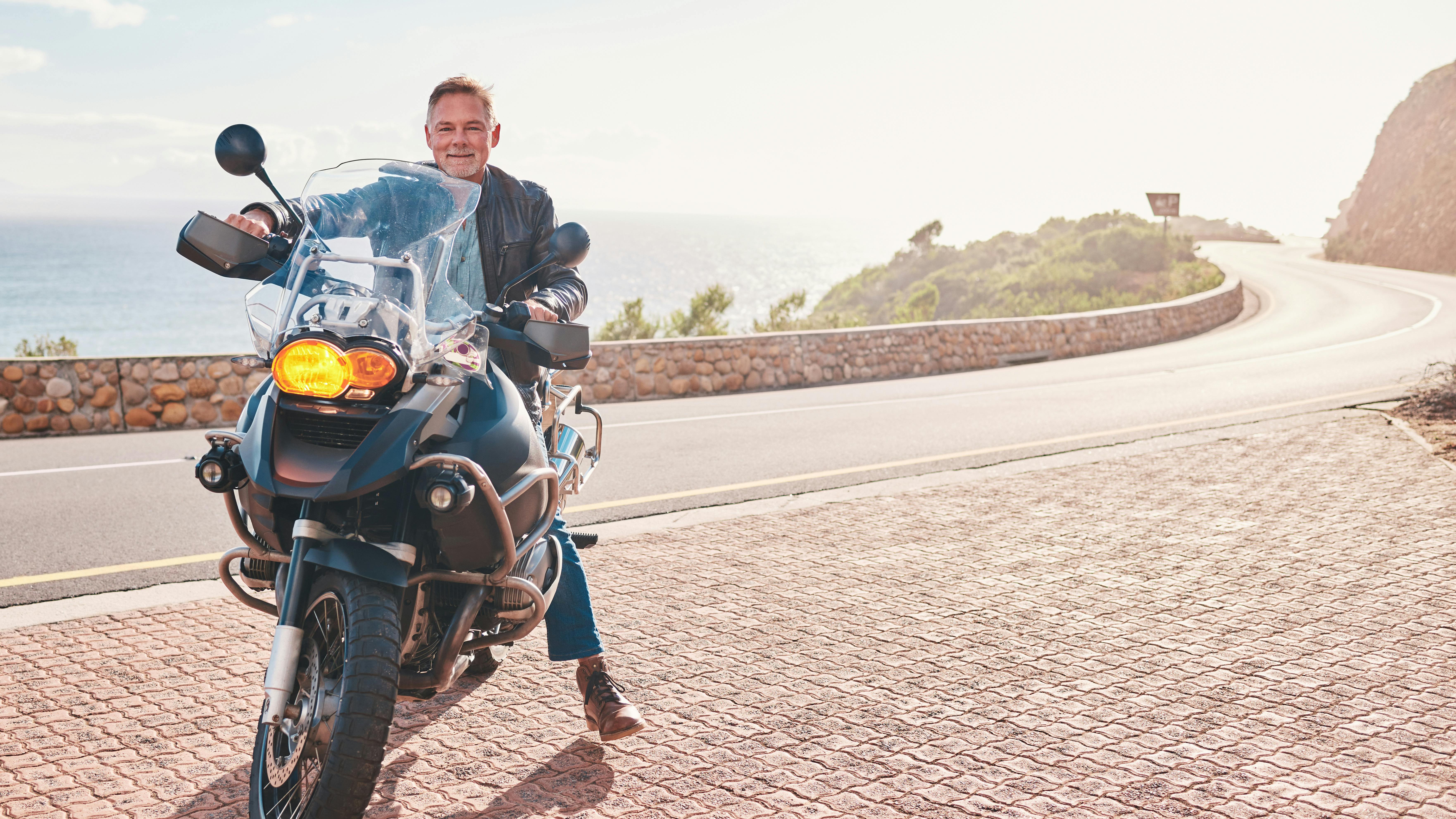Midaldrende mand holder på sin motorcykel på en bjergvej langs havet.