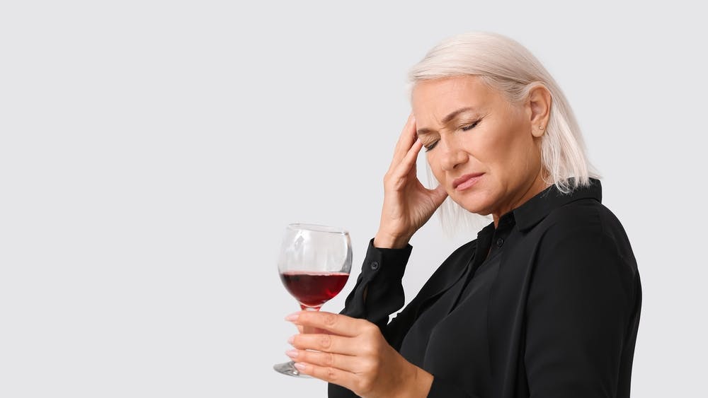 Midaldrende kvinde med et glas rødvin i hånden tager sig til hovedet.