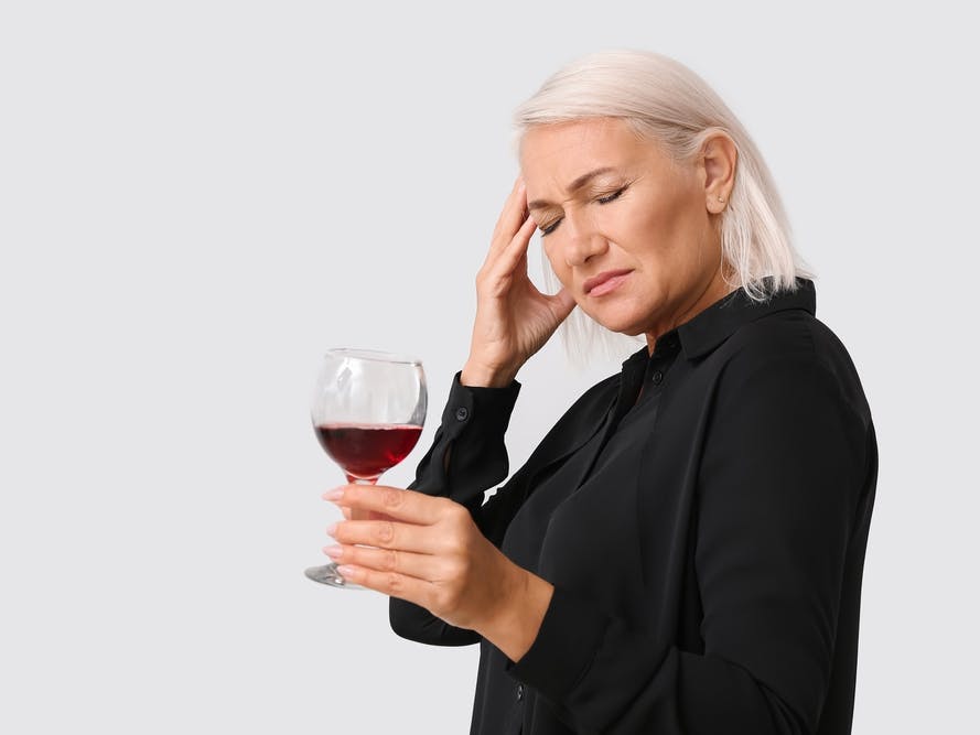 Midaldrende kvinde med et glas rødvin i hånden tager sig til hovedet.