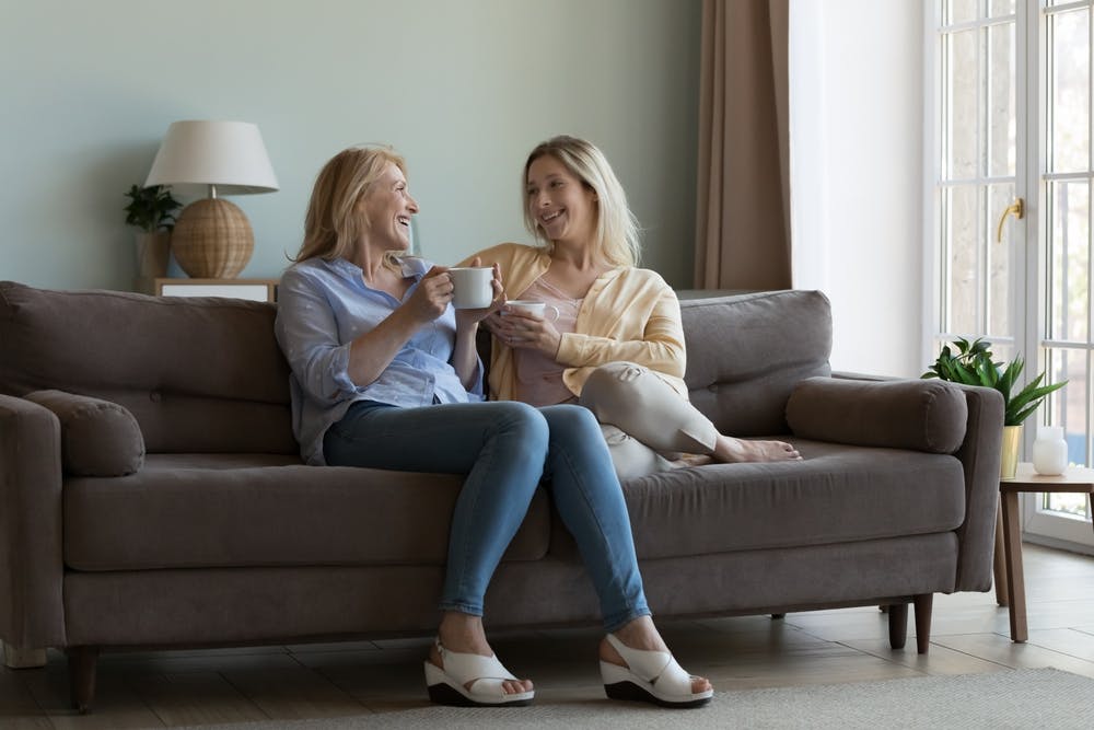To kvinder sidder i en sofa og drikker kaffe, mens de snakker og griner.