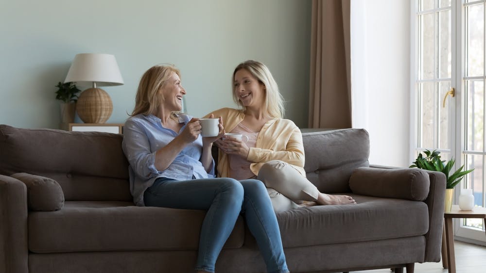 To kvinder sidder i en sofa og drikker kaffe, mens de snakker og griner.