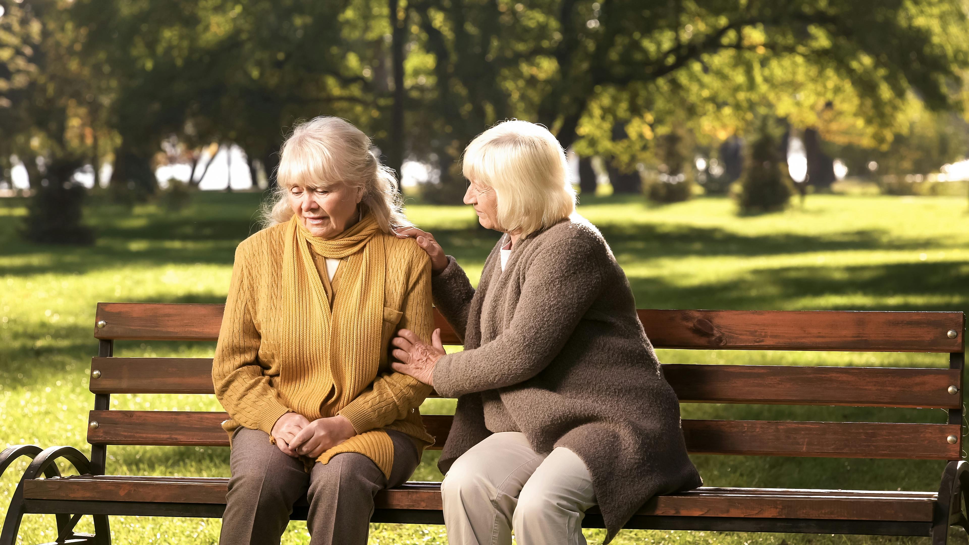 To ældre kvinder sidder på en bænk, og den ene lægger en trøstende hånd på den andens arm