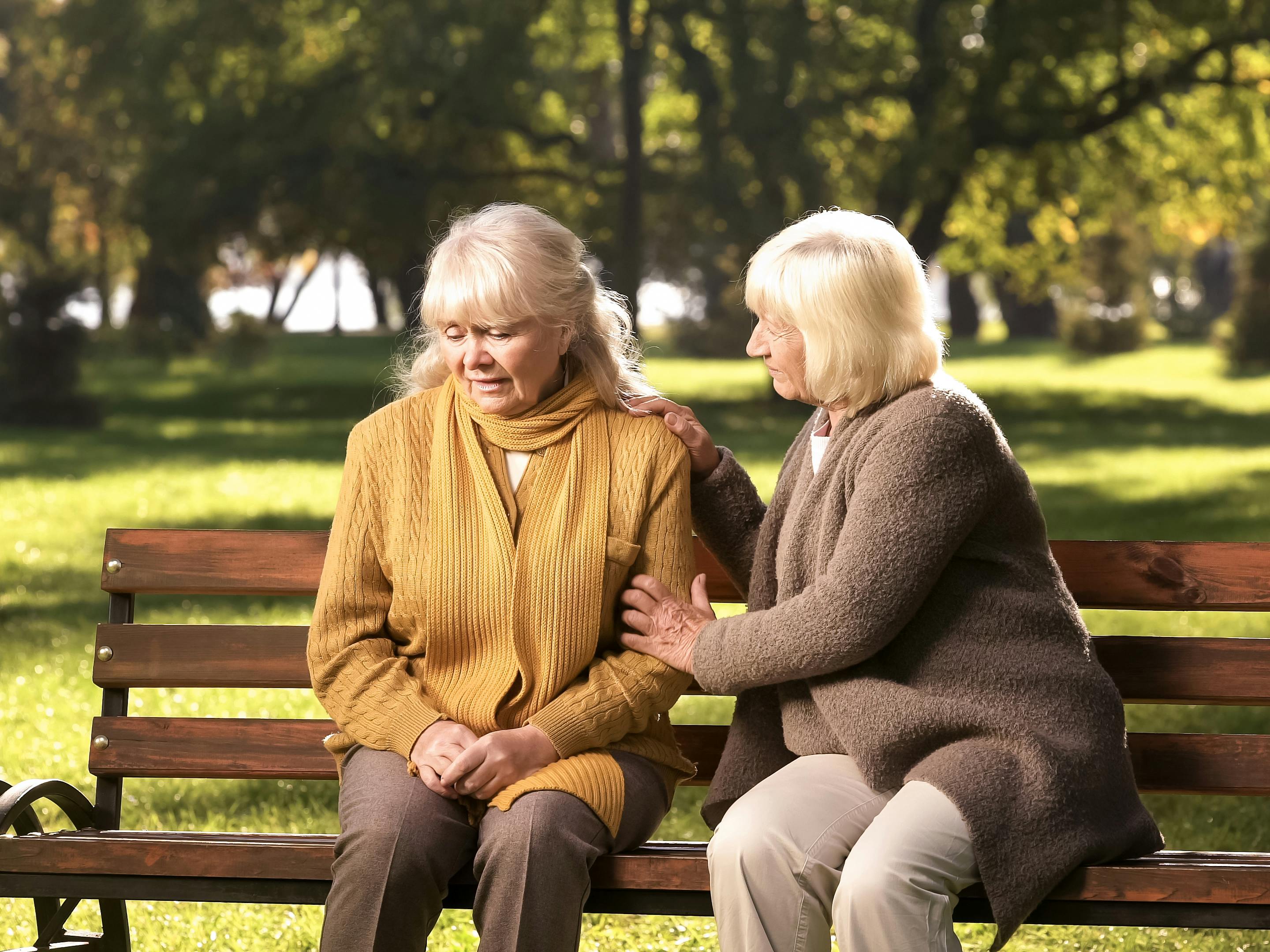 To ældre kvinder sidder på en bænk, og den ene lægger en trøstende hånd på den andens arm