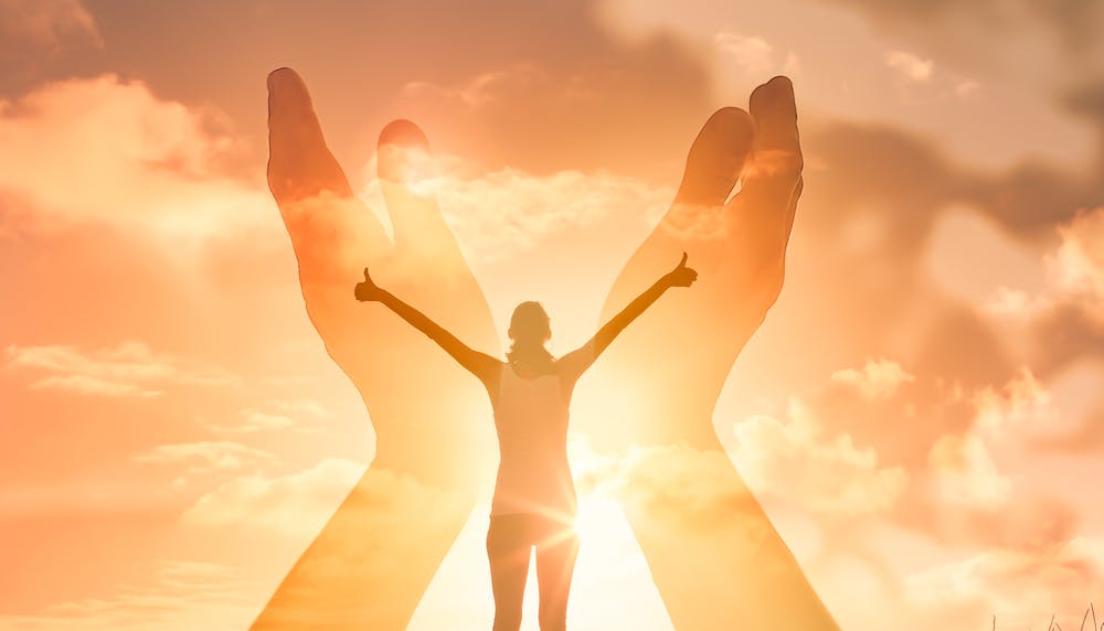 Kvinde står i gult sollys og rækker armene op i himlen. Hun er favnet af to andre hænder
