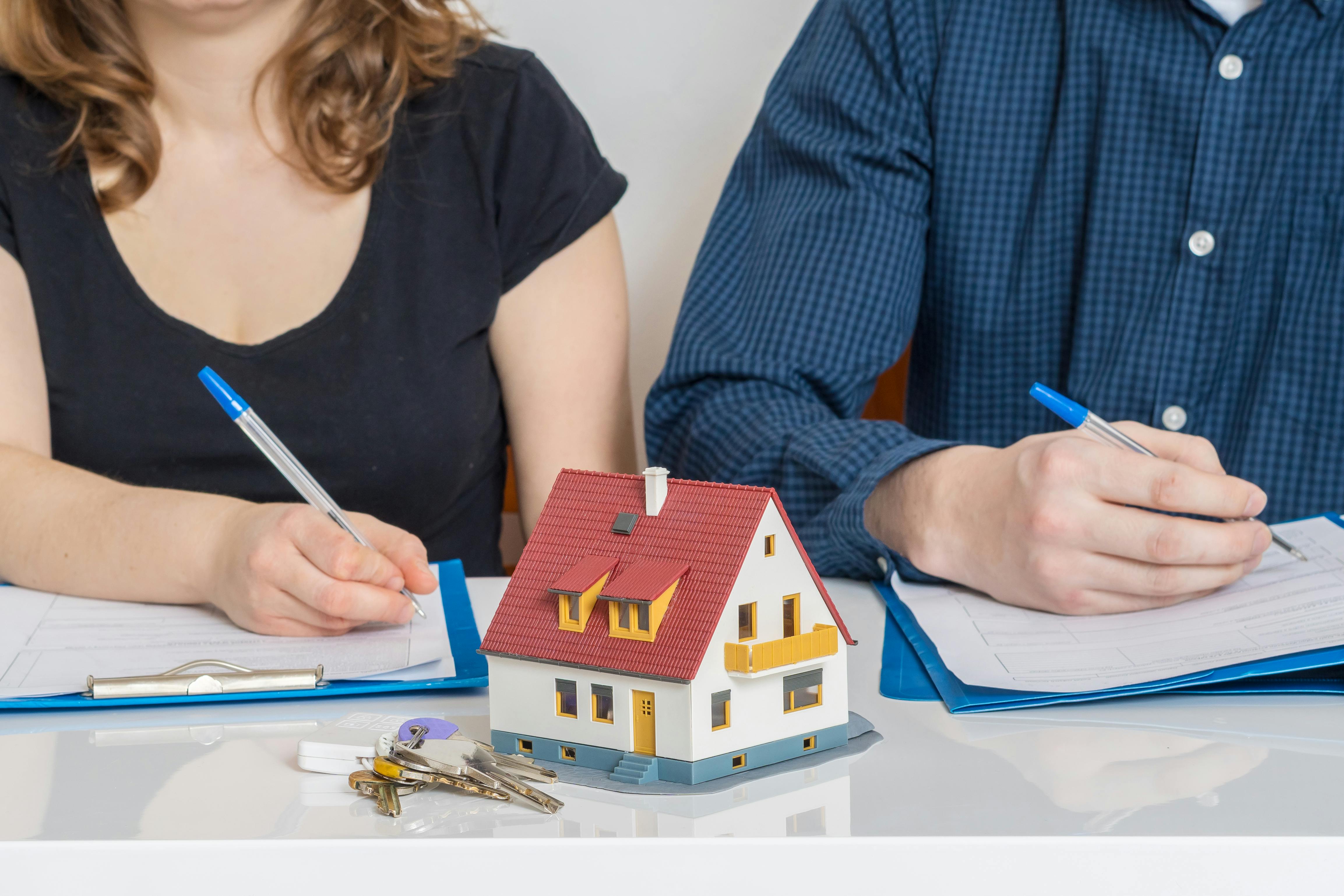Par underskriver hver sine papirer med nøgler og en model af et hus stående foran sig på bordet.