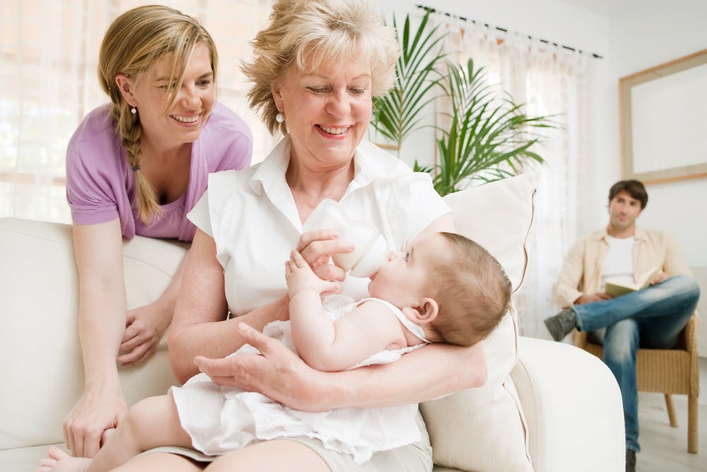 Mormor sidder med et lille barn i favnen, mens mor ser glædestrålende til. I baggrunden sidder faren og ser trist ud.