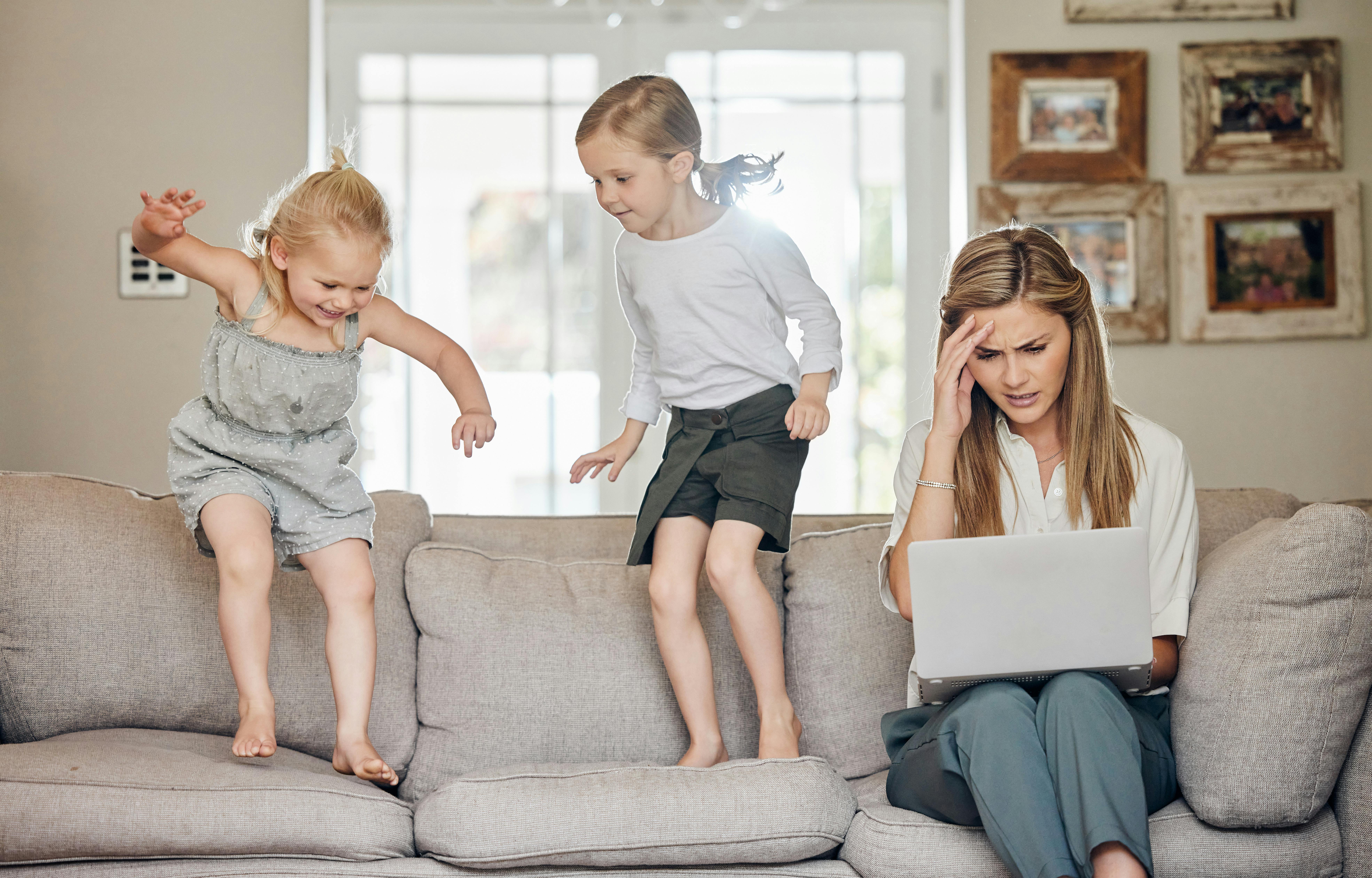 Ung mor sidder med sin bærbare computer og ser stresset ud, mens hendes to børn hopper i sofaen.