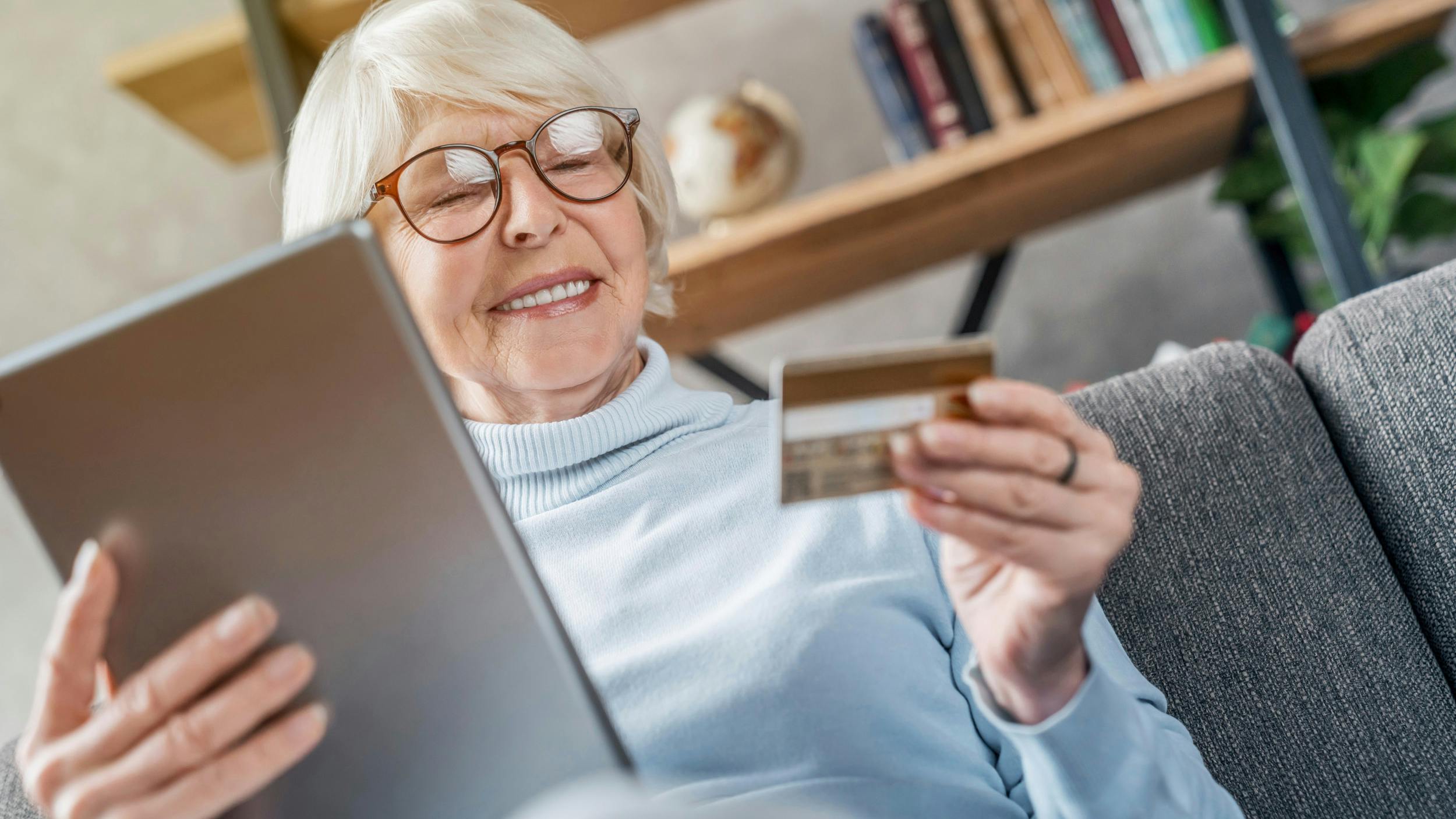 Ældre kvinde ser glad på sit kreditkort og har sin iPad i den anden hånd klar til at shoppe på nettet.