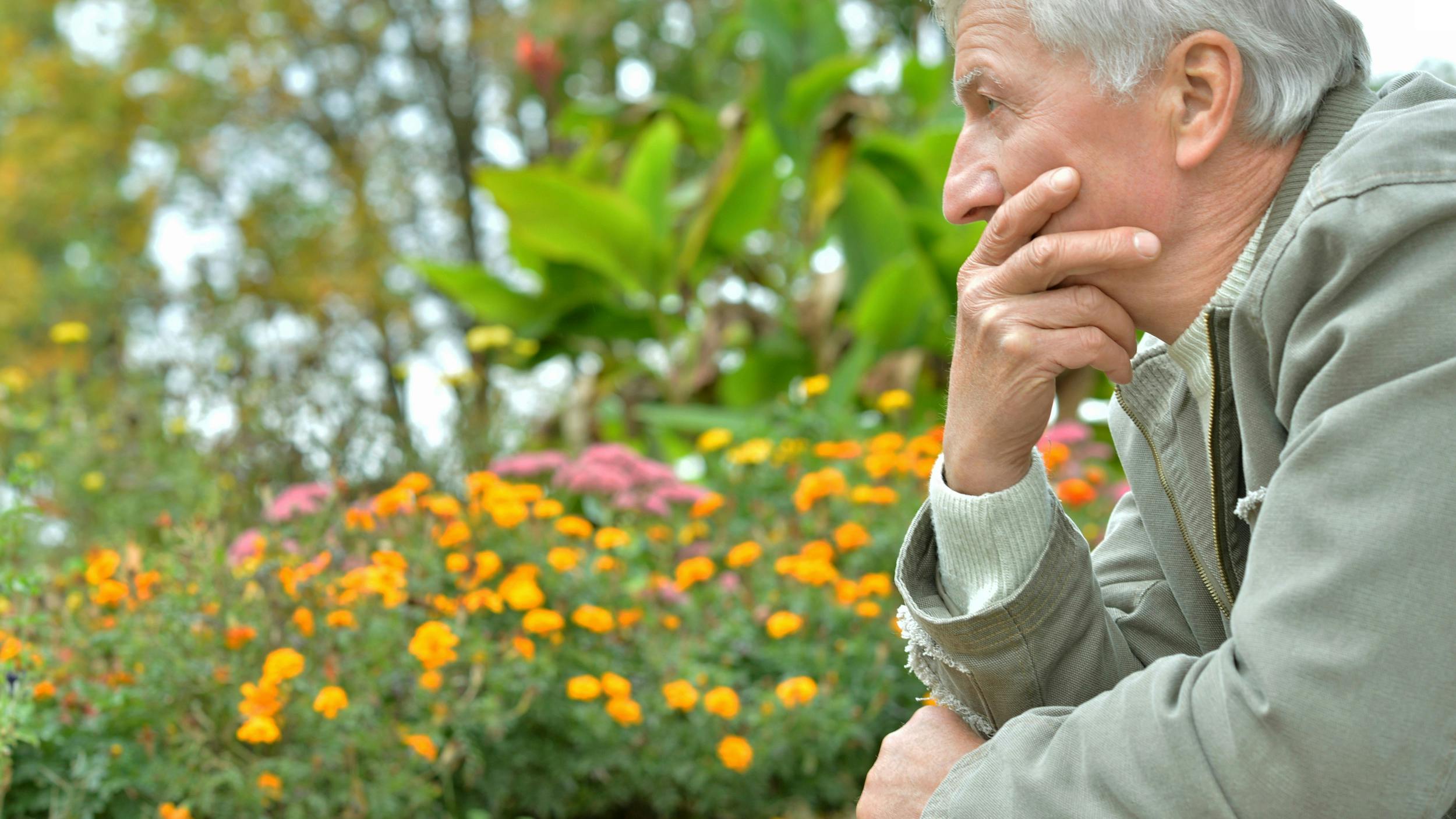 Ældre mand ser trist og eftertænksom frem for sig. Der er farverige blomster i baggrunden.
