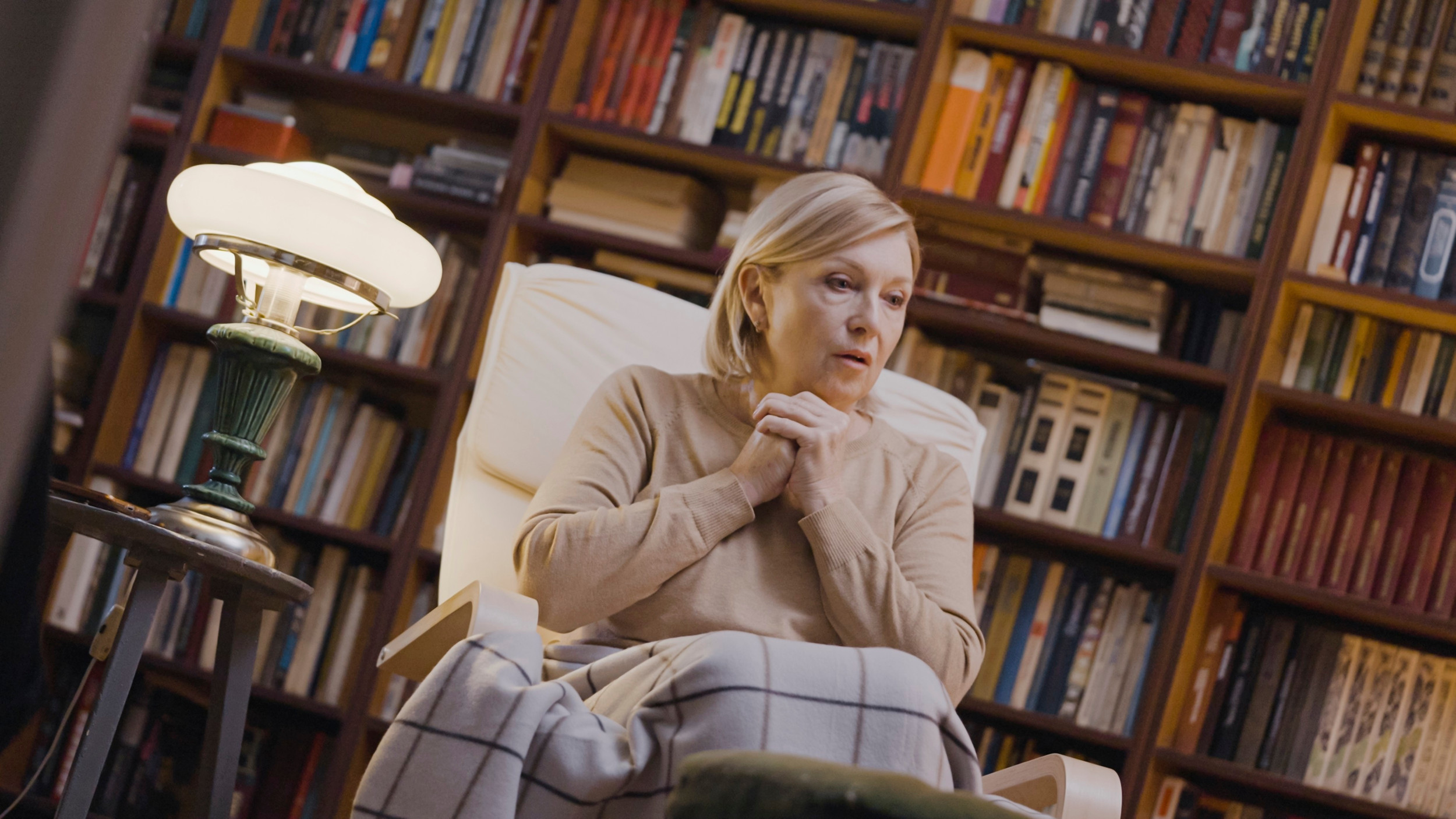 Bekymret ældre kvinde sidder alene i sit hjem omgivet af bøger.