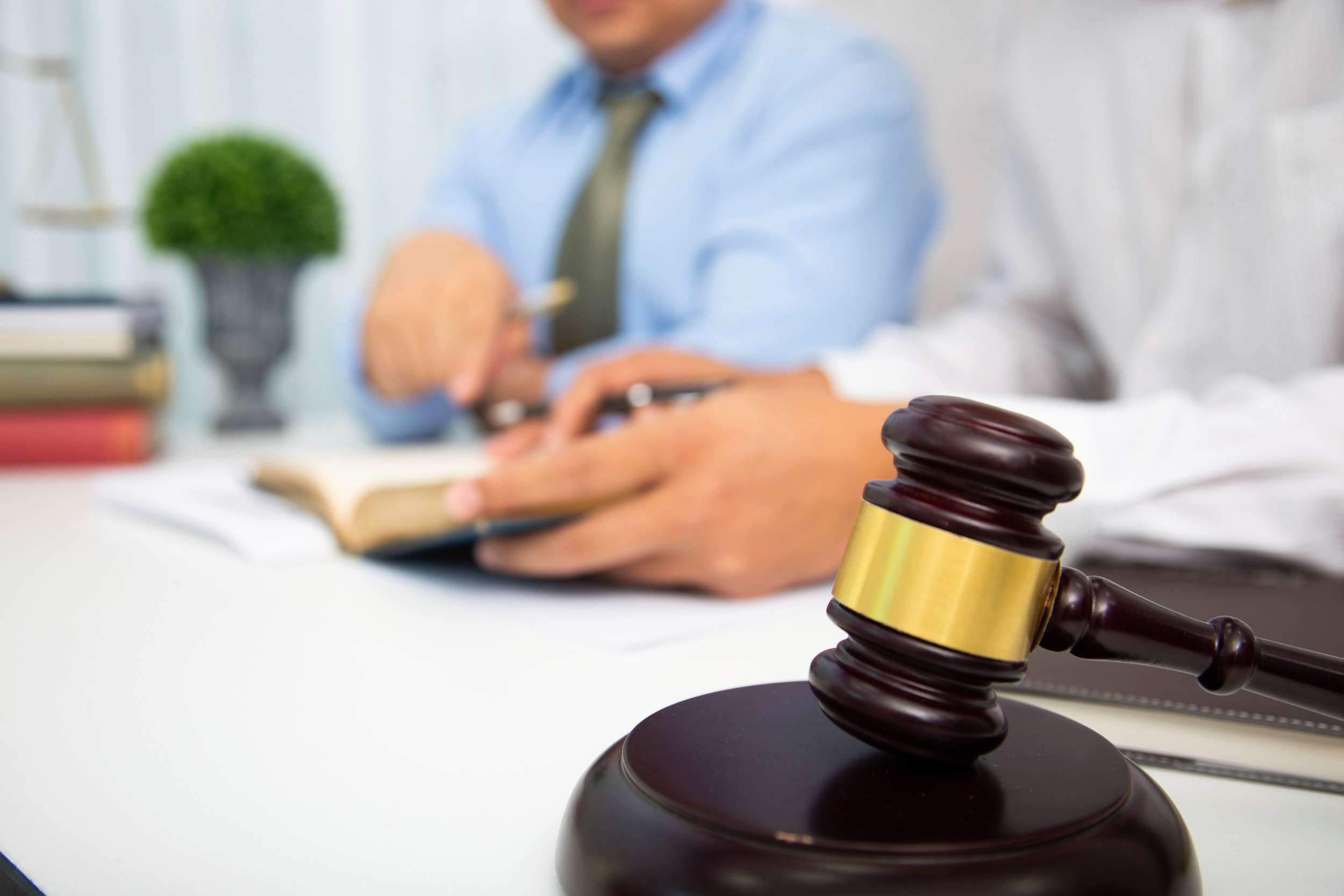 I forgrunden er der en dommerhammer og bagved sidder to mænd ved et skrivebord og bladrer i en bog.