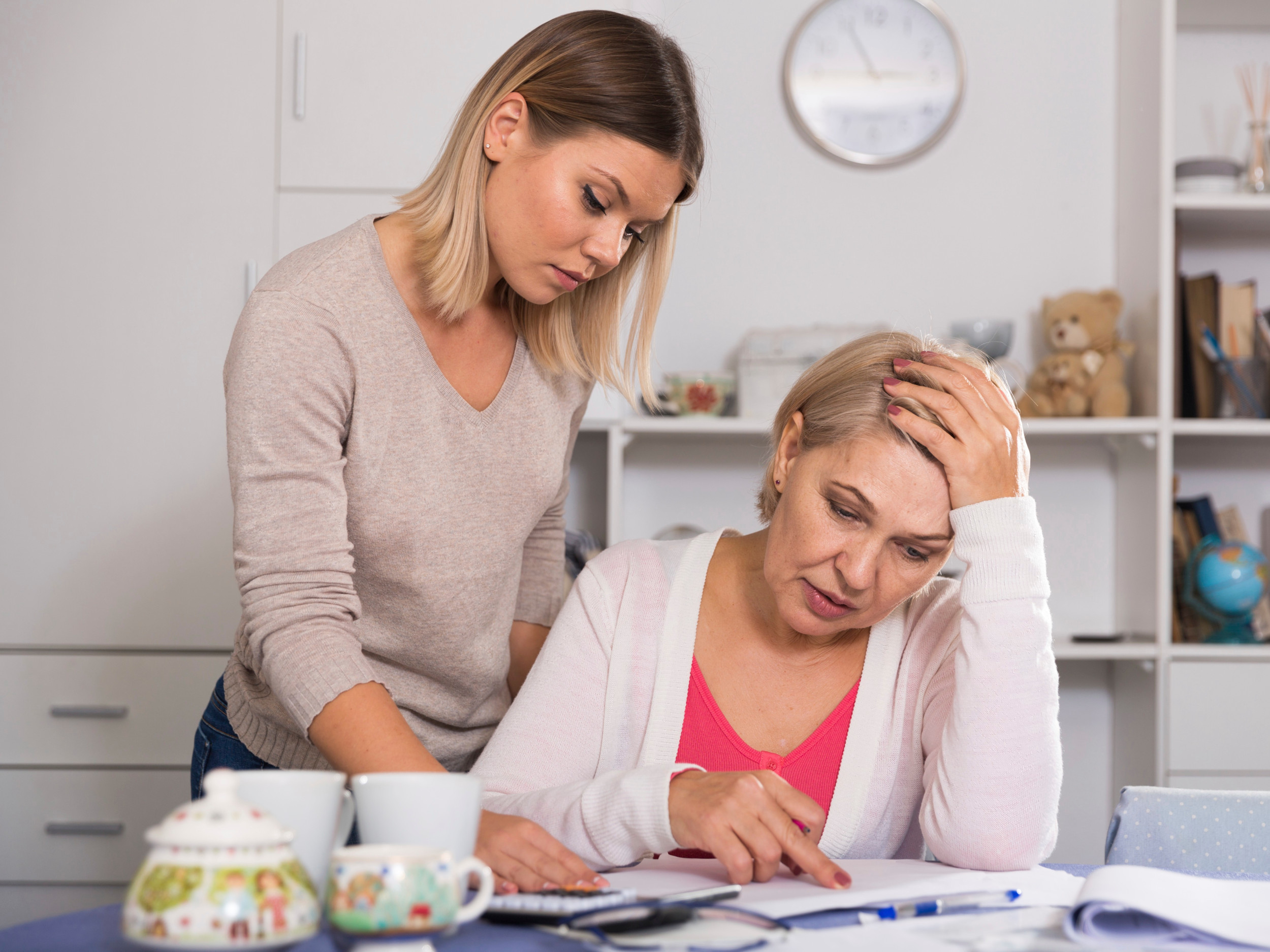 En kvinde sidder og ser opgivende ned i nogle papirer, mens hendes voksne datter står bag hende og ser på. 