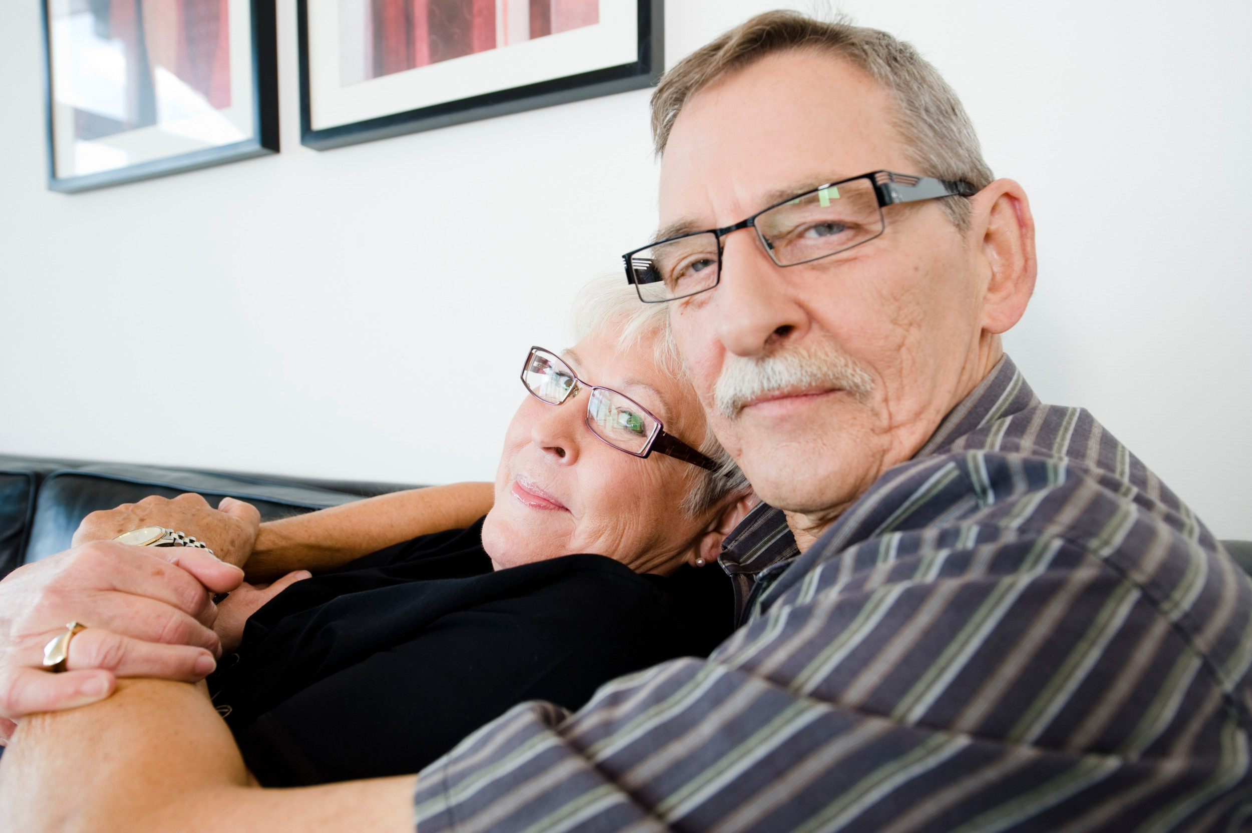 Ældre par sidder sammen i en sofa, hvor han ser lidt skeptisk ud, men hun ser forelsket på ham.