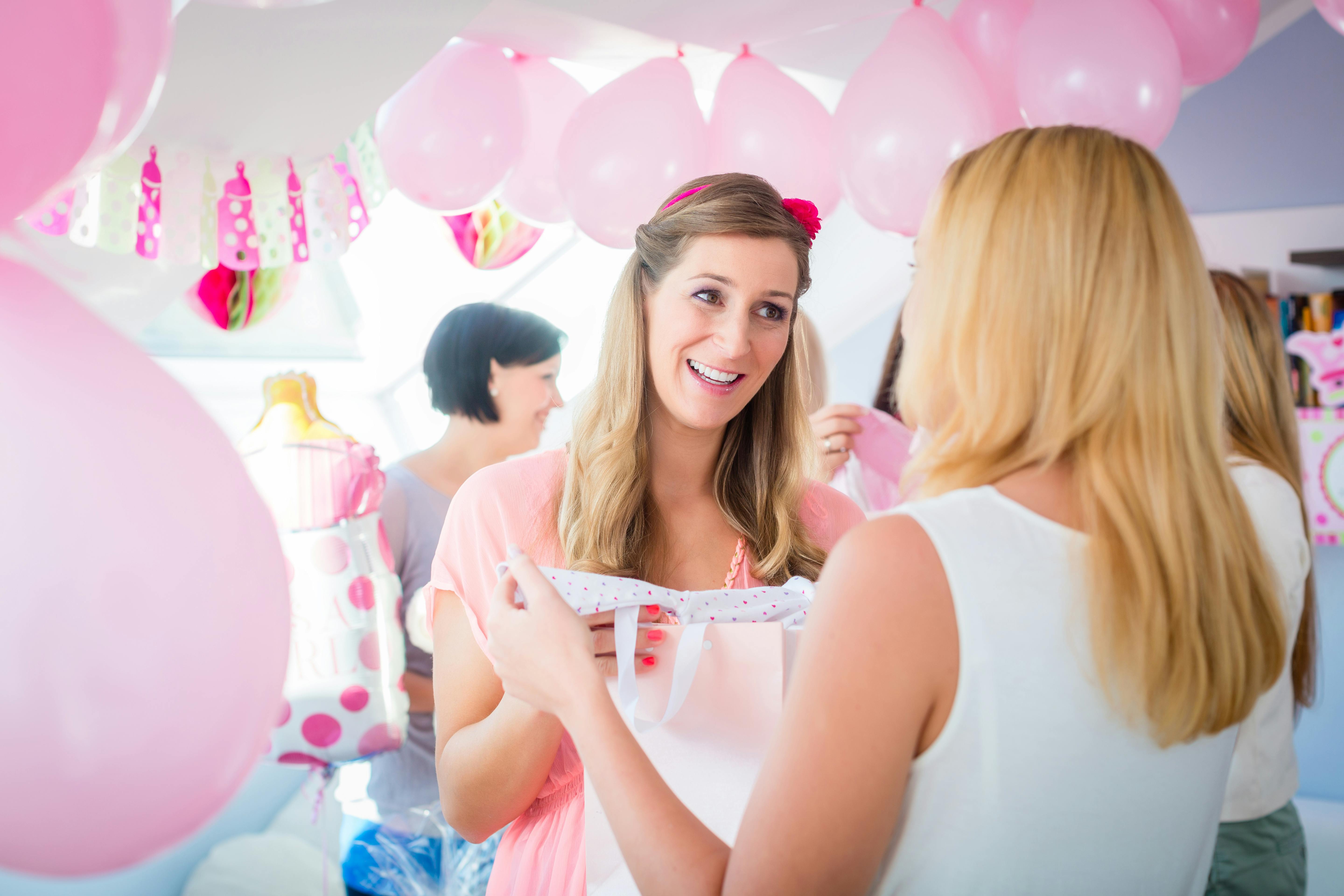 Gravid kvinde holder noget babytøj frem, som hun har fået til babyshower. Rummet er pyntet med lyserøde balloner, og i baggrunden står flere veninder.