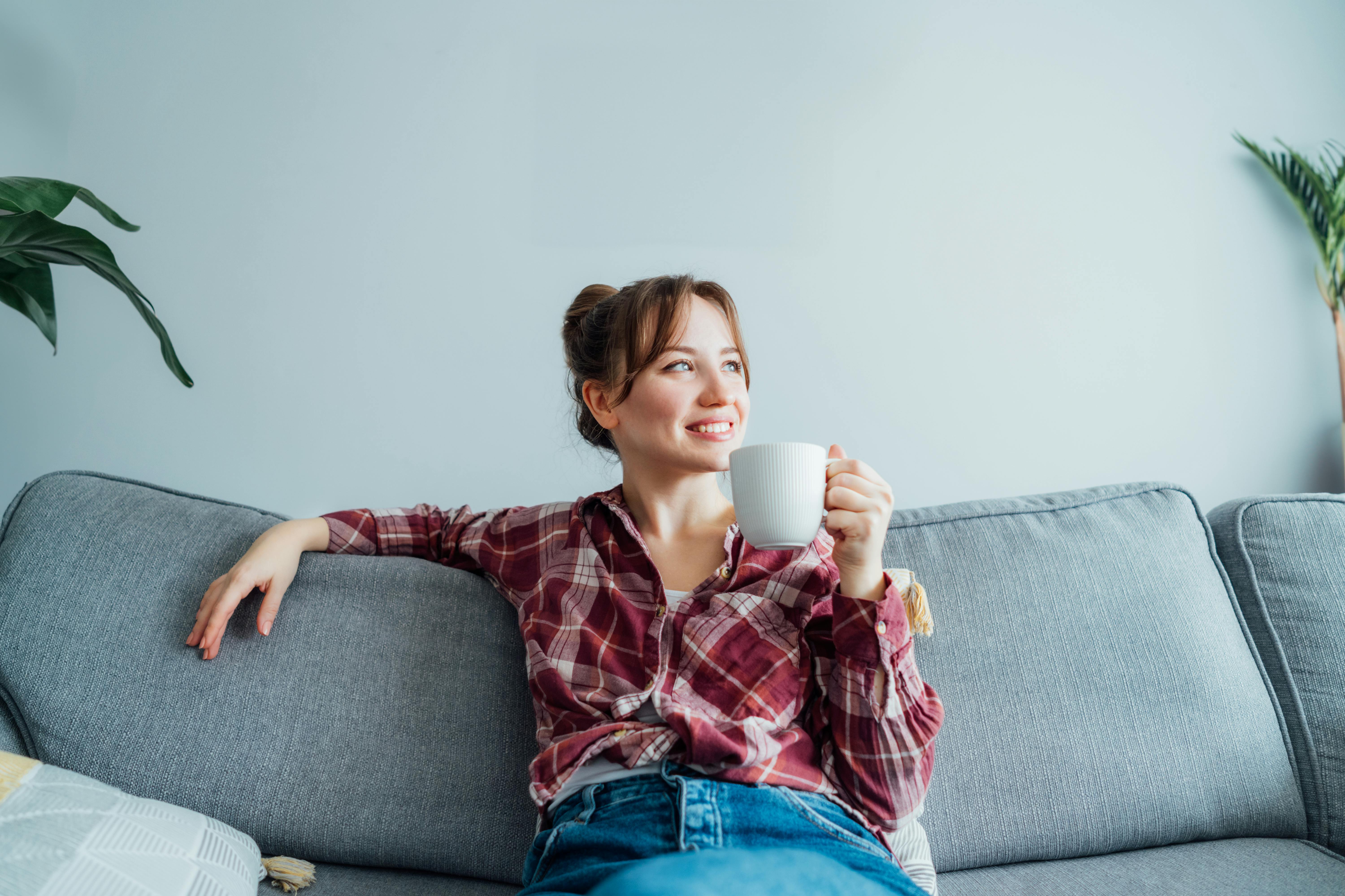 Kvinde sidder i en sofa med en kop kaffe og ser drømmende frem for sig med et stort smil om munden.