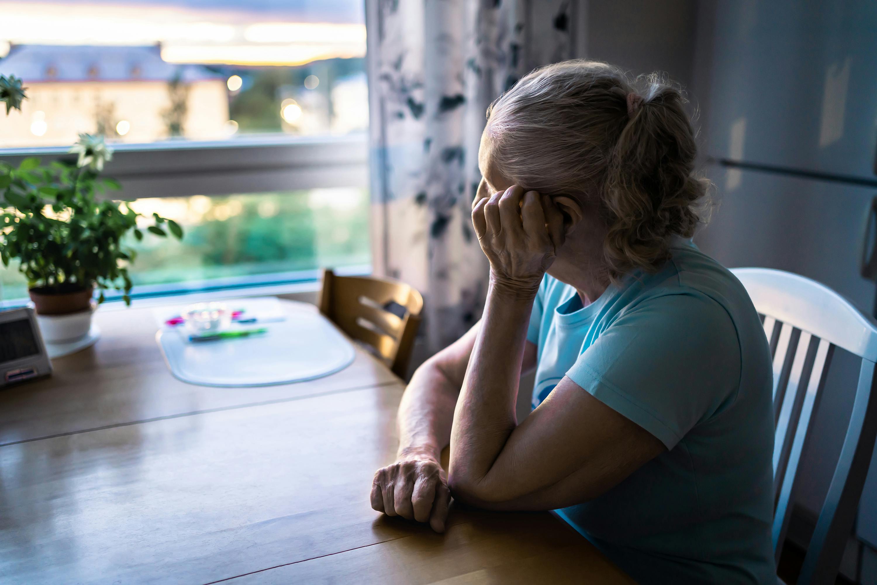 Ensom ældre kvinde sidder ved sit spisebord og ser ud af vinduet.