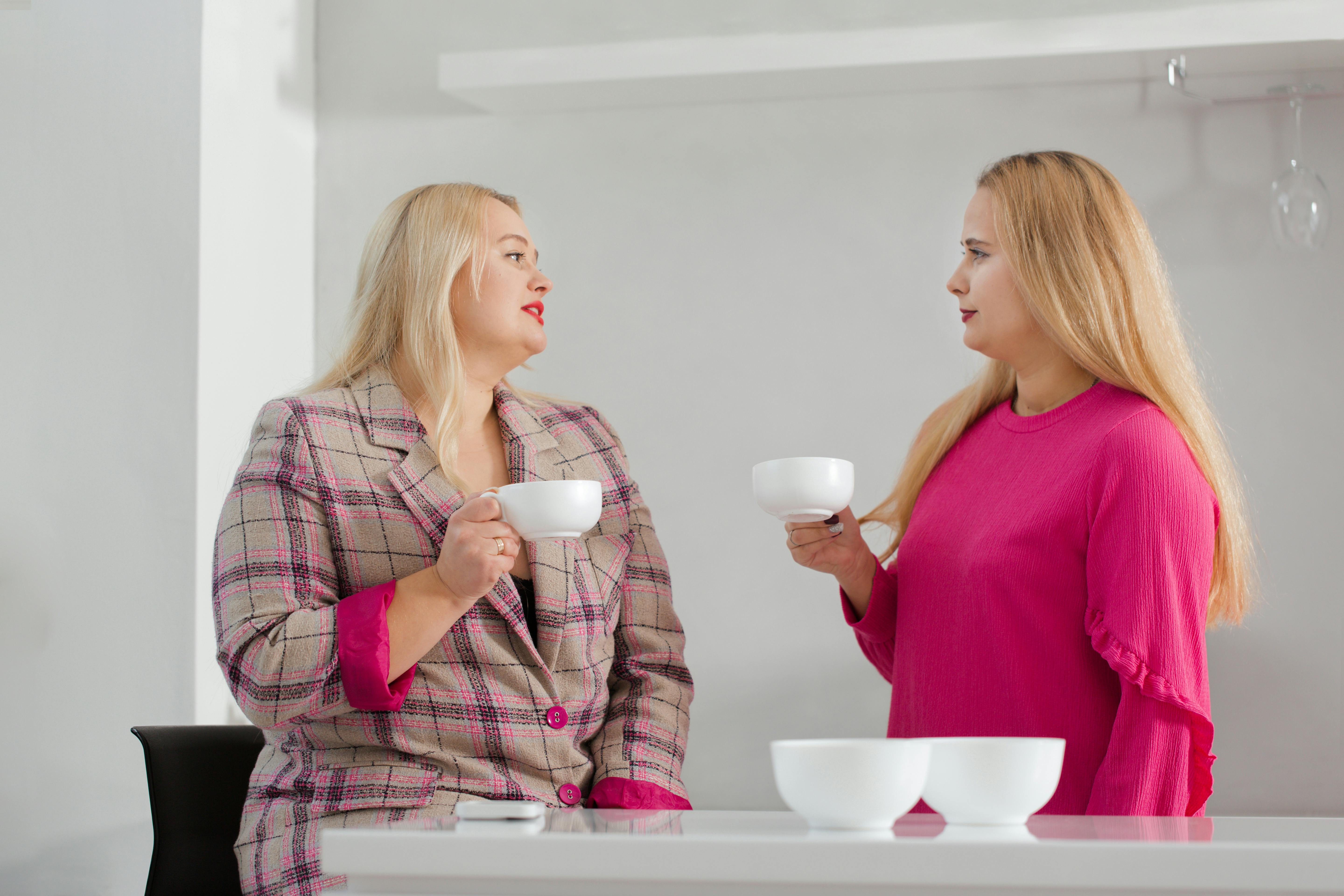 To søstre står i køkkenet med hver sin kop kaffe. Den ene af dem er overvægtig.